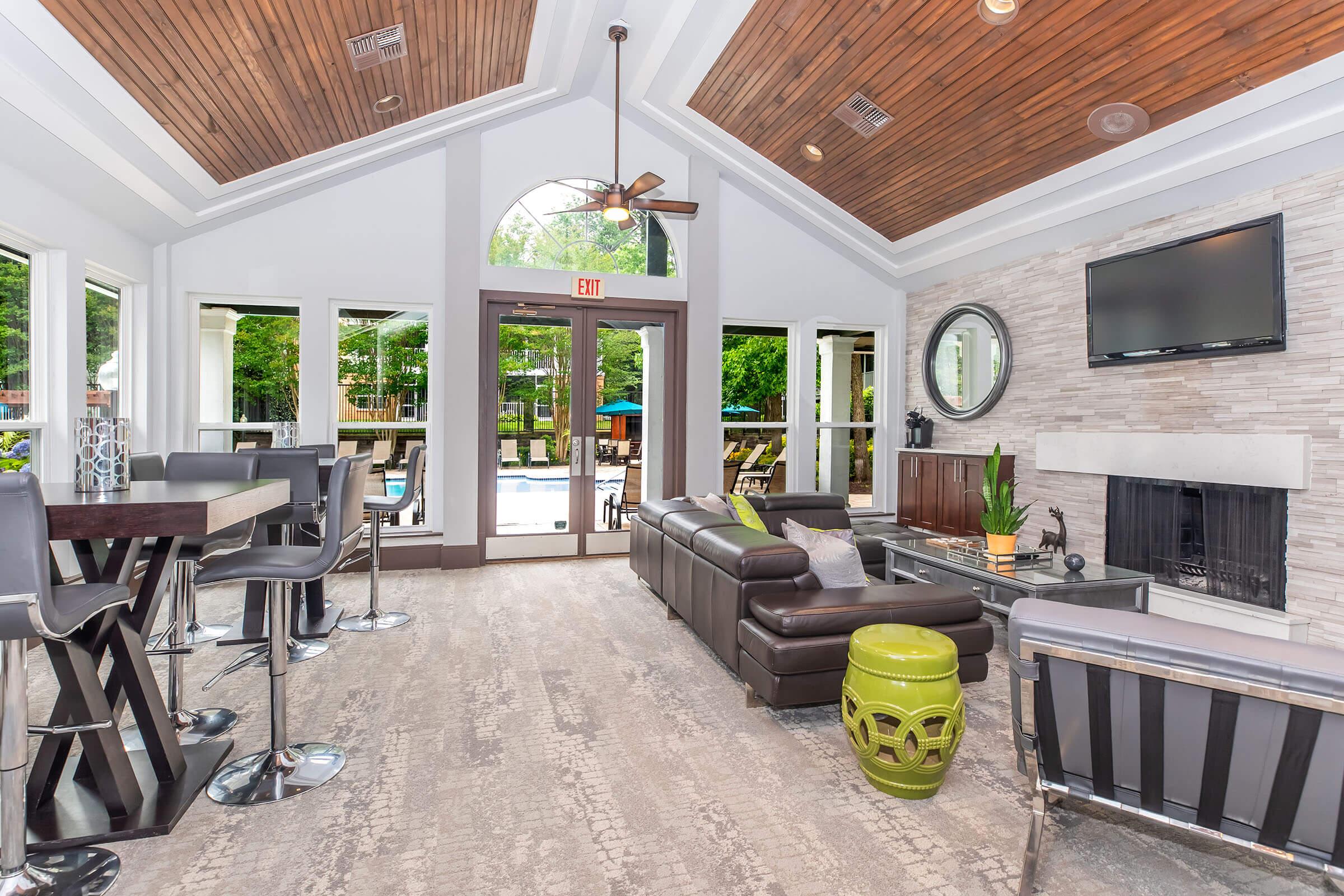 Interior of a modern lounge area featuring large windows with views of a pool, a sleek leather sofa, and stylish bar stools. The ceiling has wooden beams, and there is a stone fireplace and a flat-screen TV on the wall. Potted plants and decorative items add a fresh touch to the space.