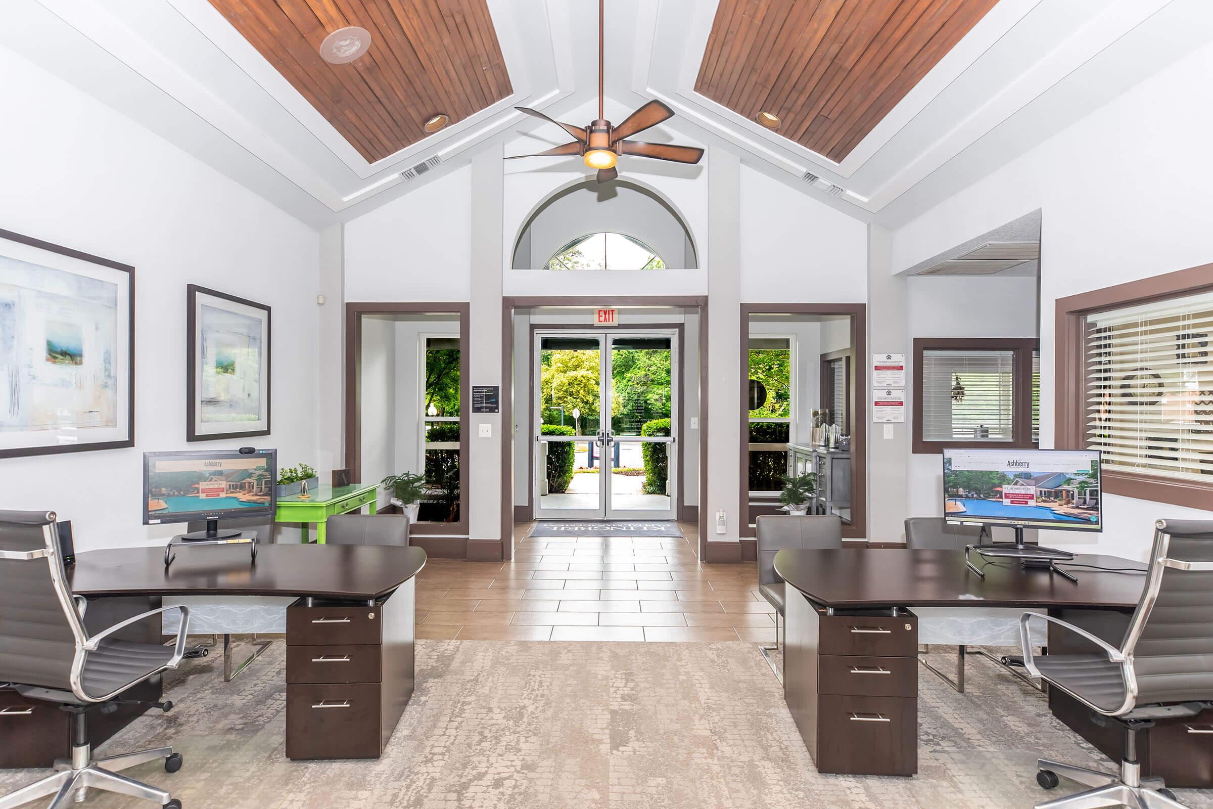 A modern office space featuring two desks with computers, stylish ceiling fans, and an open entrance leading to a well-lit outdoor area. The decor includes framed artwork and a welcoming atmosphere with greenery visible through the large windows.