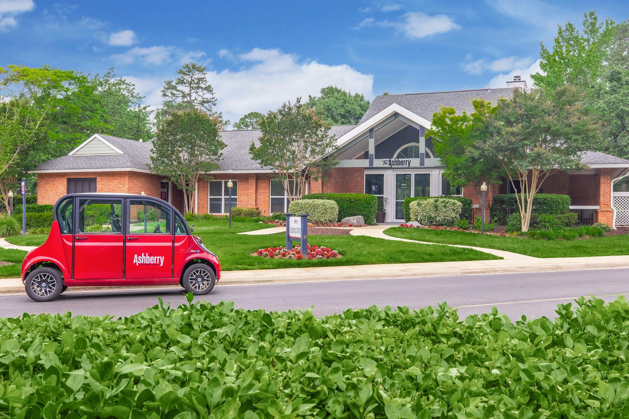 A small red electric vehicle parked in front of a modern brick building with large windows. The building features landscaping with flowers and trees, and a sign displaying "Ashberry." Bright blue skies with fluffy clouds complete the scene.