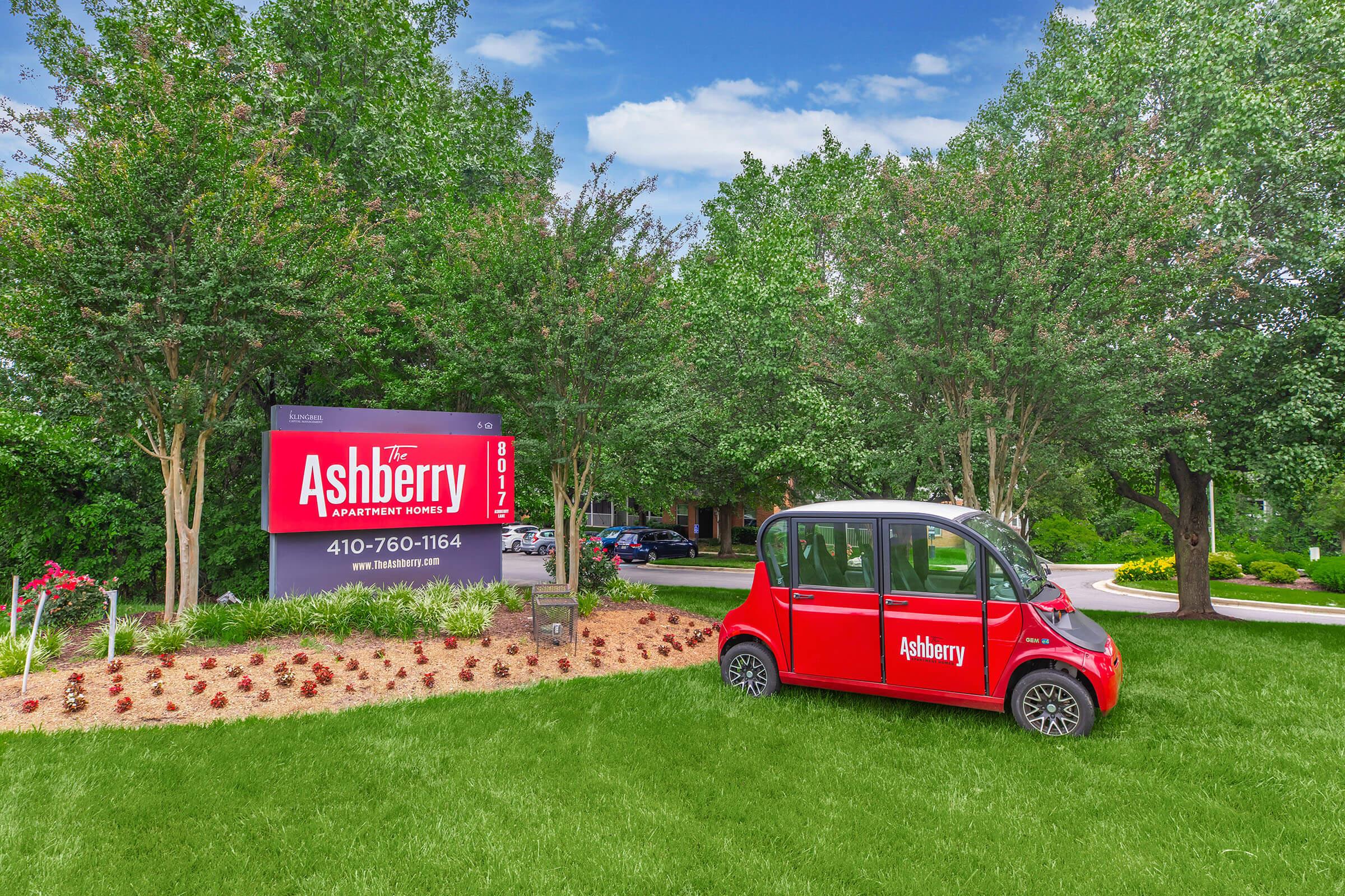 A red electric vehicle is parked in front of a large sign for The Ashberry apartment complex. The sign features the name "The Ashberry" prominently, alongside contact information. Lush green grass and well-maintained landscaping frame the scene, with trees providing a natural backdrop.