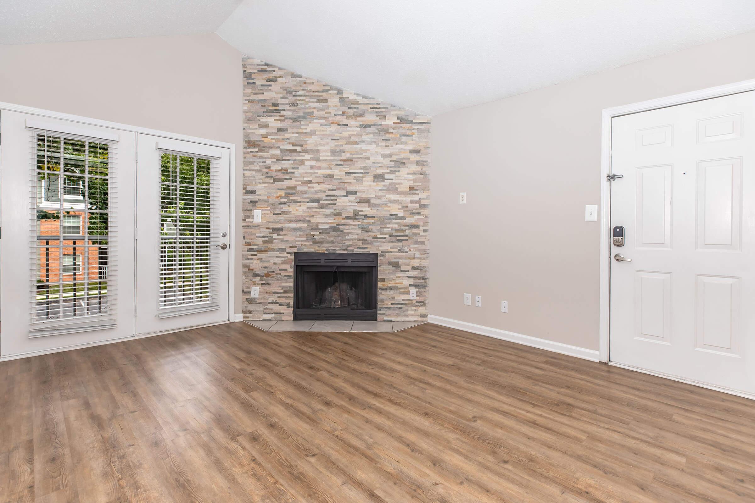 Living room interior featuring a faux stone fireplace against a textured wall, hardwood-style flooring, and large windows with white blinds. The entrance door is visible on the right, and the overall color scheme is neutral with light walls.