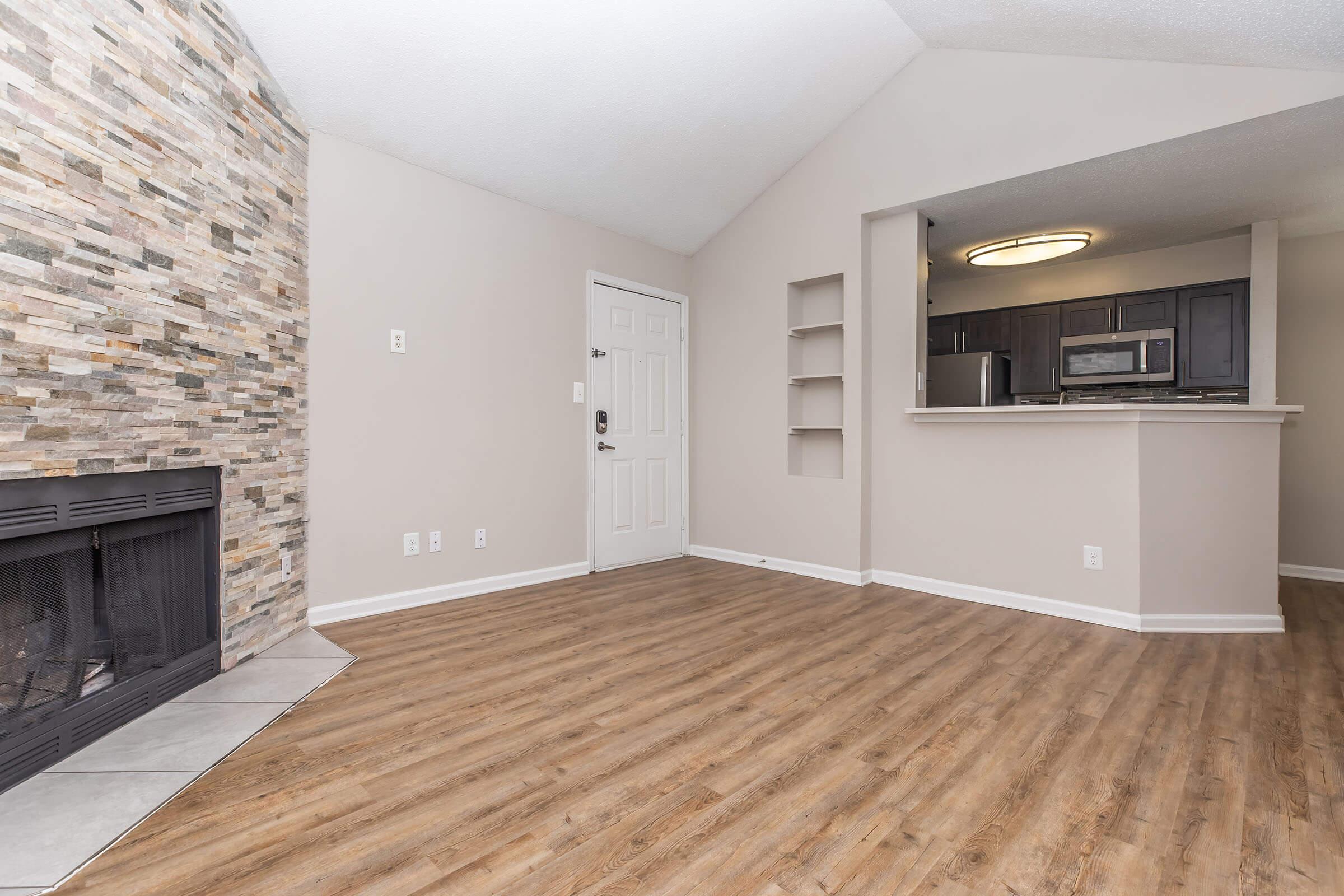 Spacious living area featuring a stone accent wall and a fireplace. The room is painted in neutral tones with hardwood-style flooring. An open layout leads to a kitchen area, which includes modern appliances, and there are built-in shelves on one wall. Natural light enhances the inviting atmosphere.