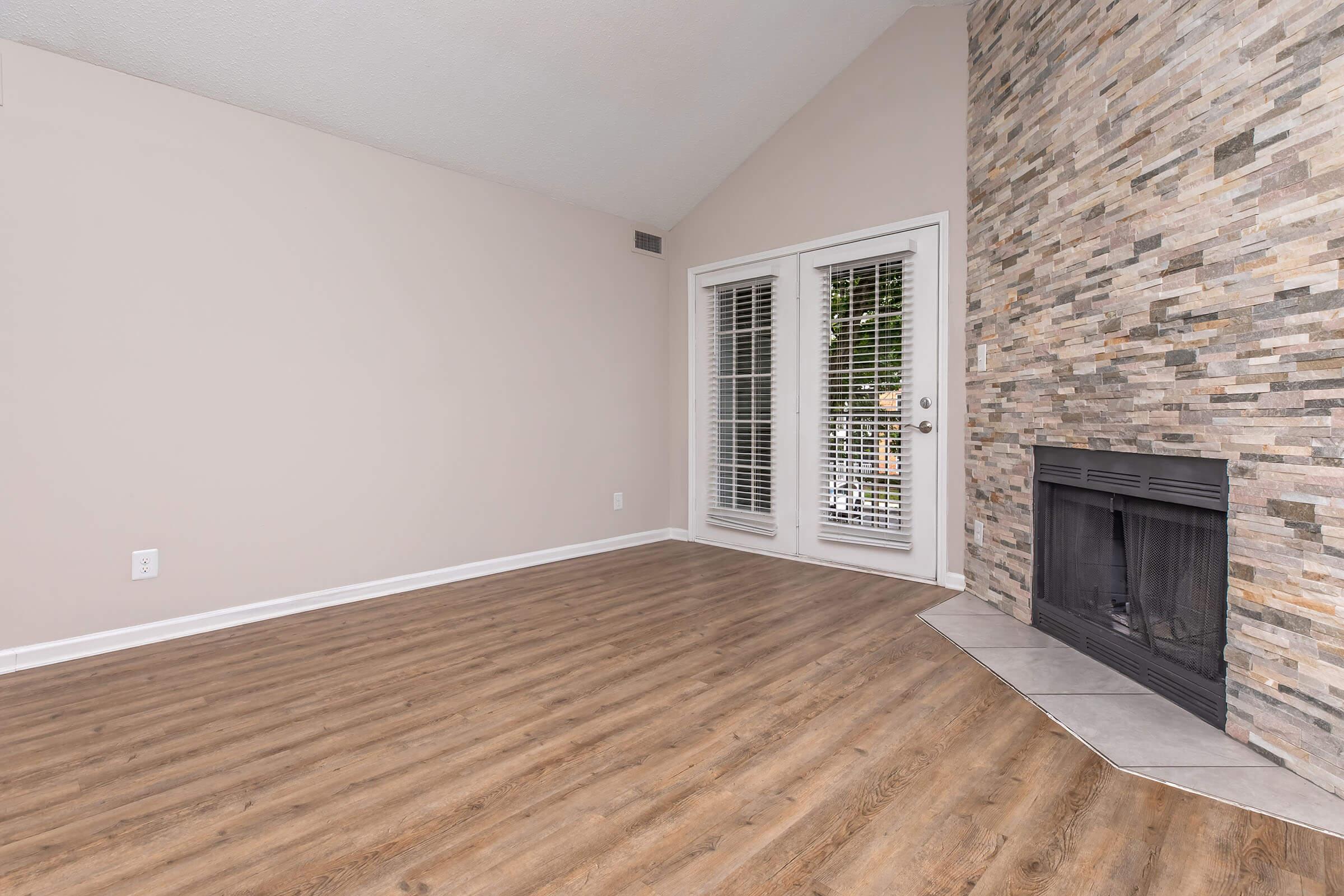 A spacious room with light-colored walls and wooden flooring. The area features a large stone fireplace against one wall and double doors with blinds leading to an outdoor space. The ceiling is vaulted, creating an open and airy feel.