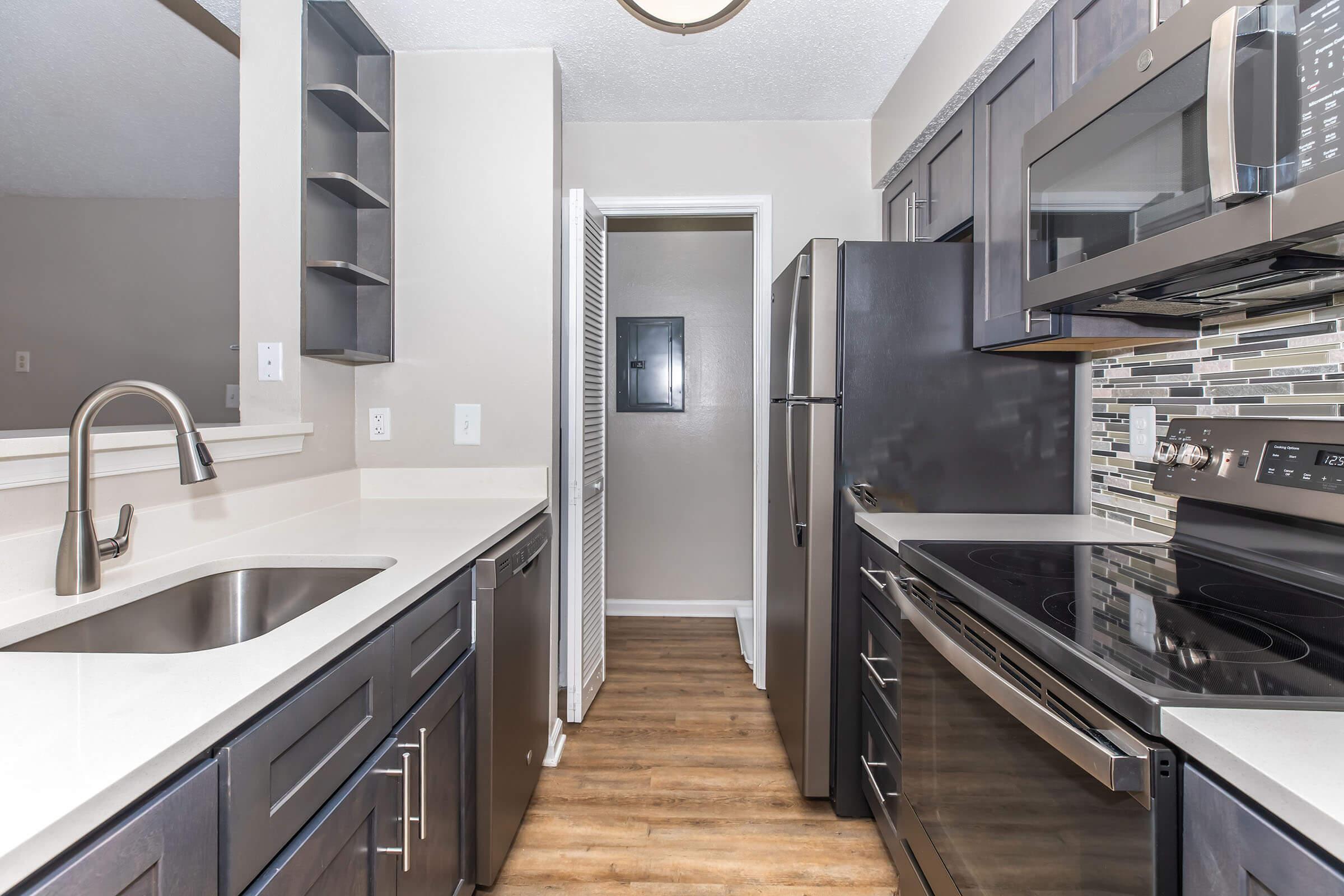 Modern kitchen featuring dark cabinetry, stainless steel appliances, and a stylish backsplash. The layout includes a double sink, built-in microwave, and an adjacent doorway leading to another room. The flooring is a warm wood tone, creating a cozy atmosphere in the space.