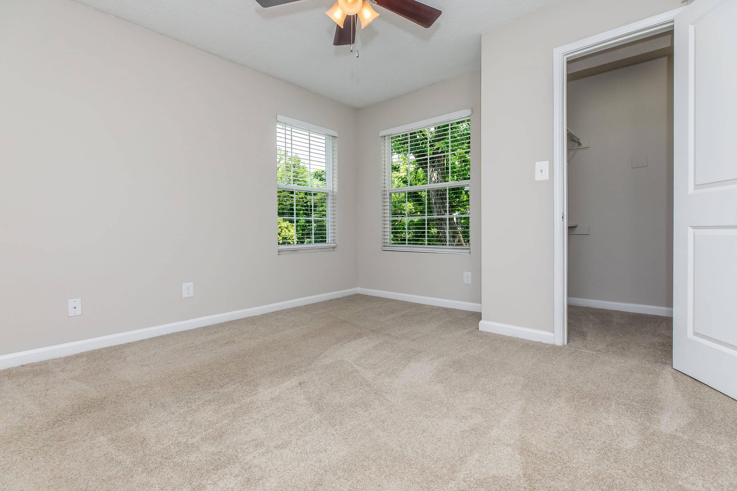 Empty room with light beige walls and carpet, featuring two windows with white blinds allowing natural light. A ceiling fan hangs from the ceiling, and a doorway leads to a small closet. The overall atmosphere is clean and bright, suitable for various purposes.