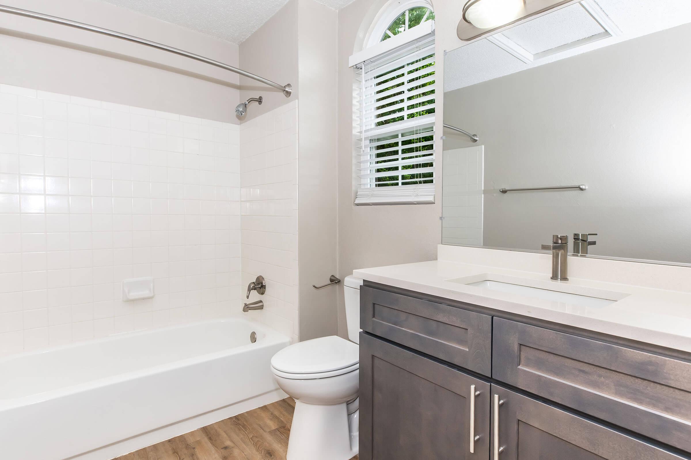 Modern bathroom featuring a white bathtub and shower combo, a white sink with a dark wood vanity, and a toilet. Natural light comes through a window with white blinds, and the walls are painted a light neutral color. The floor is a wood-like laminate.