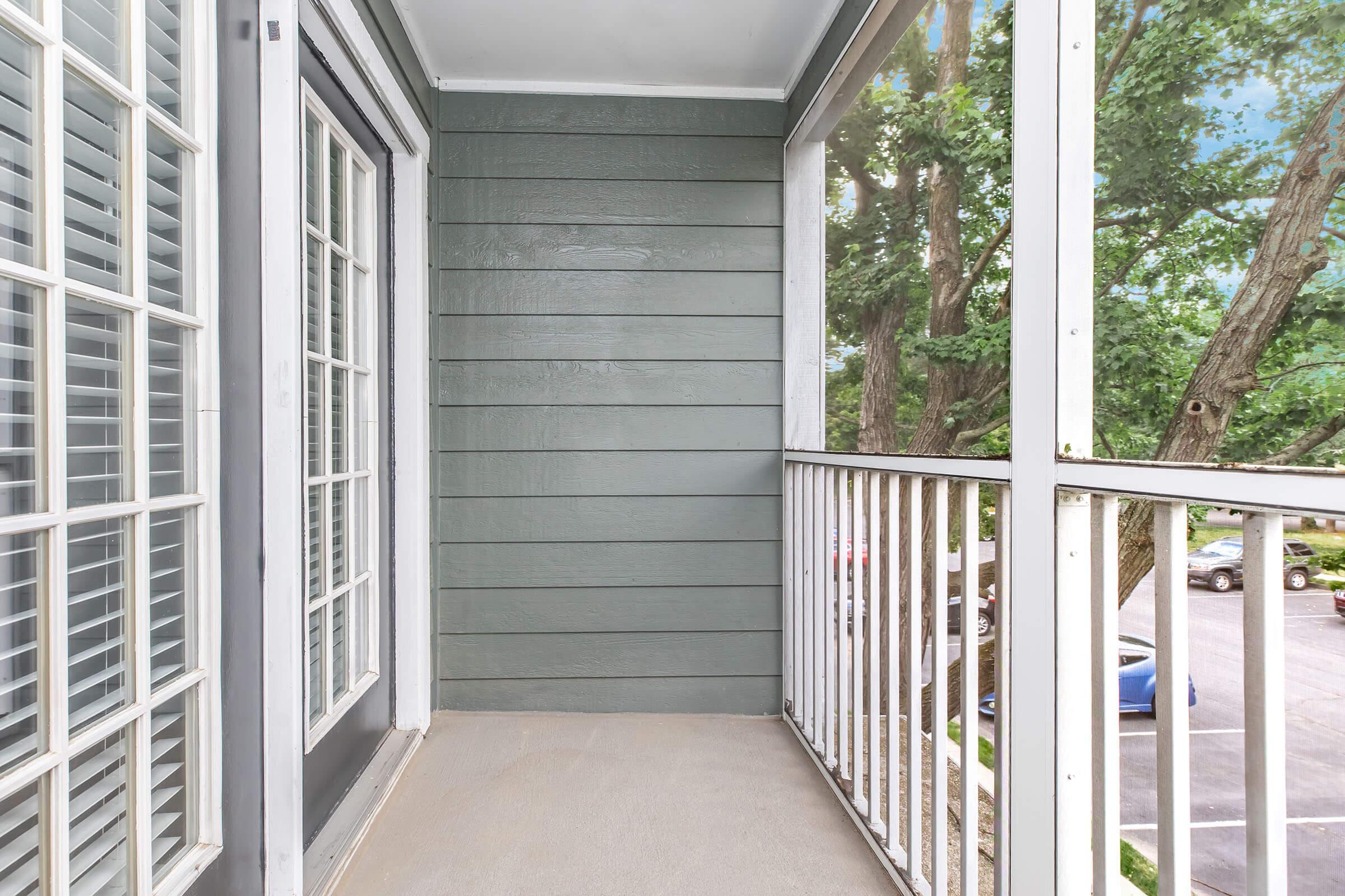 Balcony view with gray wood paneling, white railing, and large windows. The area is well-lit, showcasing trees outside and a glimpse of parked cars below.
