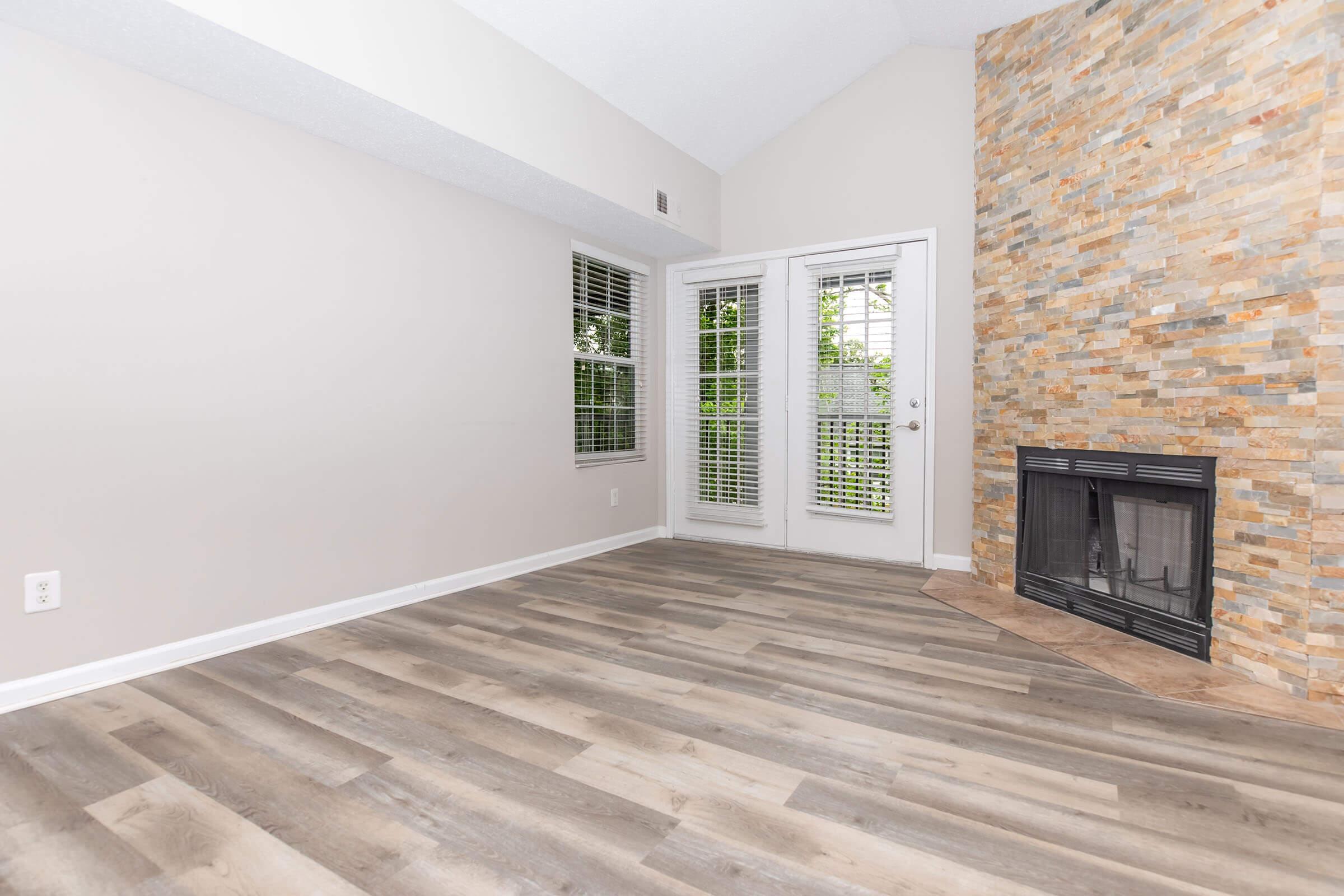 A bright, empty living room featuring light-colored walls and a stone accent wall. Large windows with white blinds allow natural light to flow in. A fireplace is situated on the right side, with hardwood floors adding warmth to the space. The room has a vaulted ceiling, creating an open and airy atmosphere.