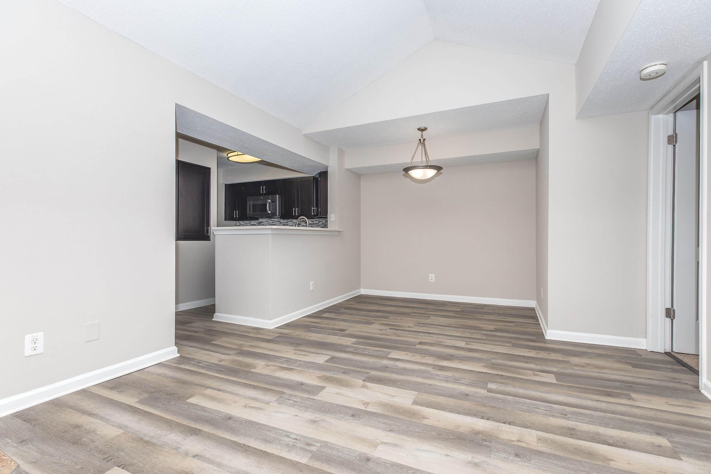 A spacious, well-lit interior of an apartment featuring a modern kitchen with dark cabinetry and a granite countertop, visible through an archway. The living area has new, light-colored flooring and a neutral wall color, complemented by a stylish light fixture overhead.