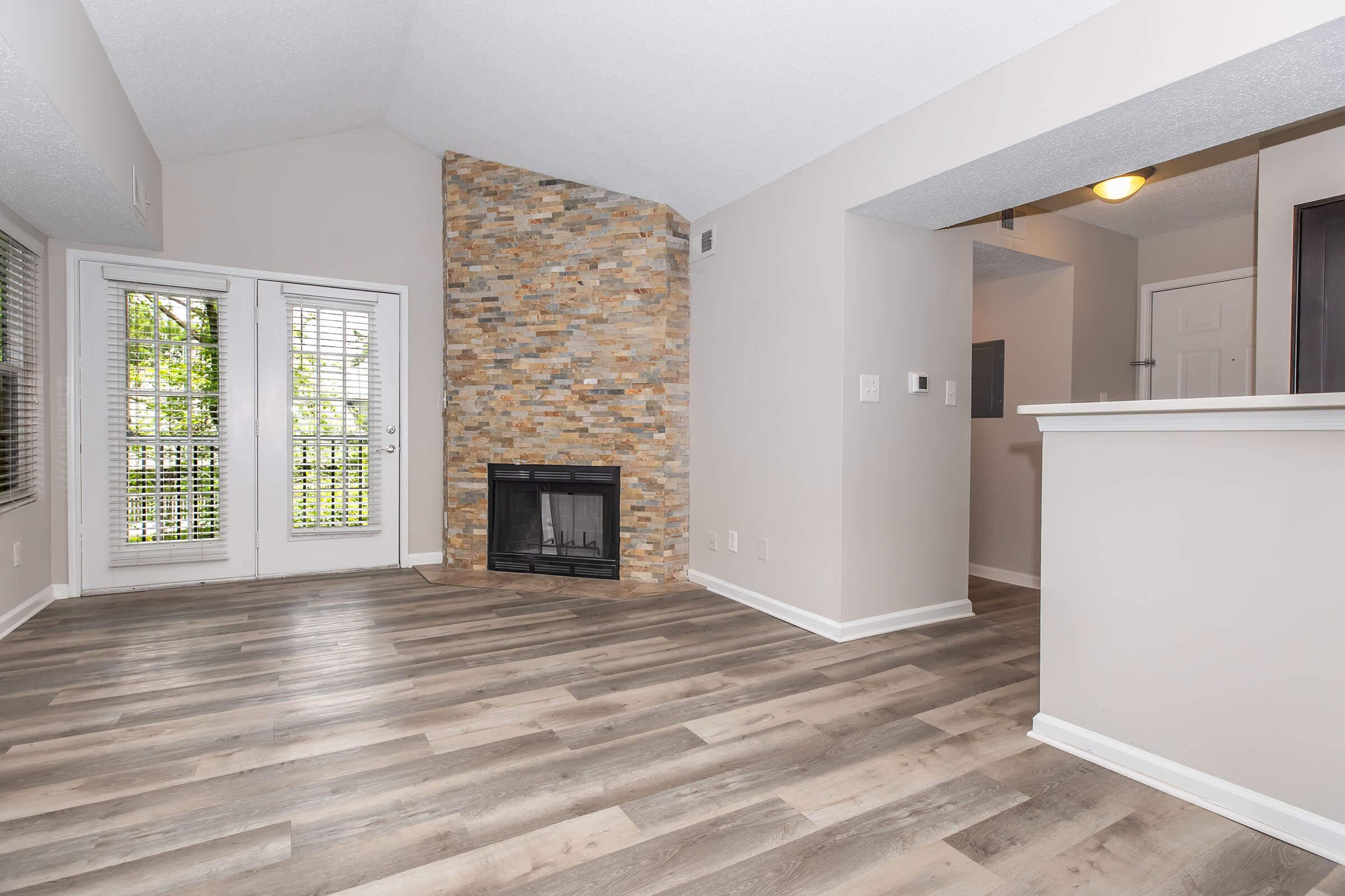 Bright, open living space featuring a stone fireplace, large windows with blinds, and modern flooring. The room has a light color scheme and leads to a kitchen area with bar seating. Ideal for a cozy, inviting atmosphere.