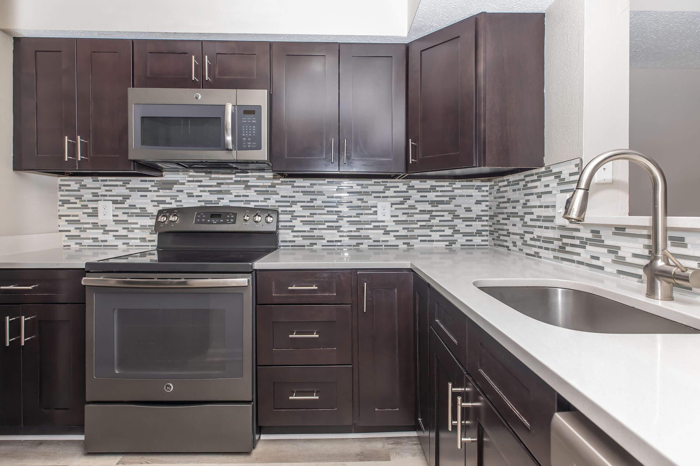 A modern kitchen featuring dark wooden cabinets, stainless steel appliances including an oven, microwave, and sink. The countertop is white with a sleek finish, and the backsplash is composed of mosaic tiles in shades of gray and blue, creating an elegant and contemporary look.
