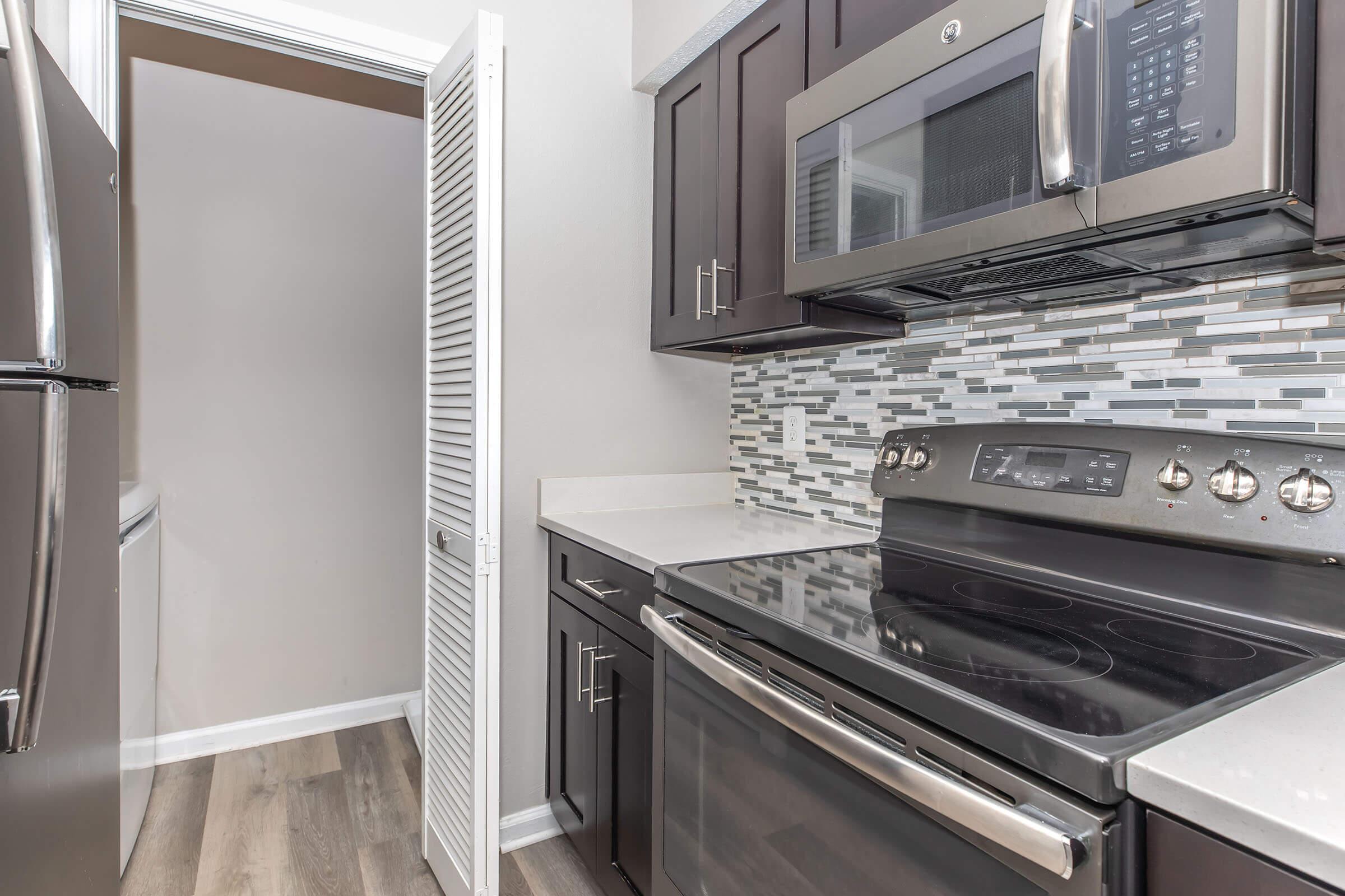 A modern kitchen featuring dark cabinetry, stainless steel appliances, including a microwave and stove, and a decorative tile backsplash. The design includes a refrigerator and an open doorway leading to another space, with light-colored countertops and wooden flooring.
