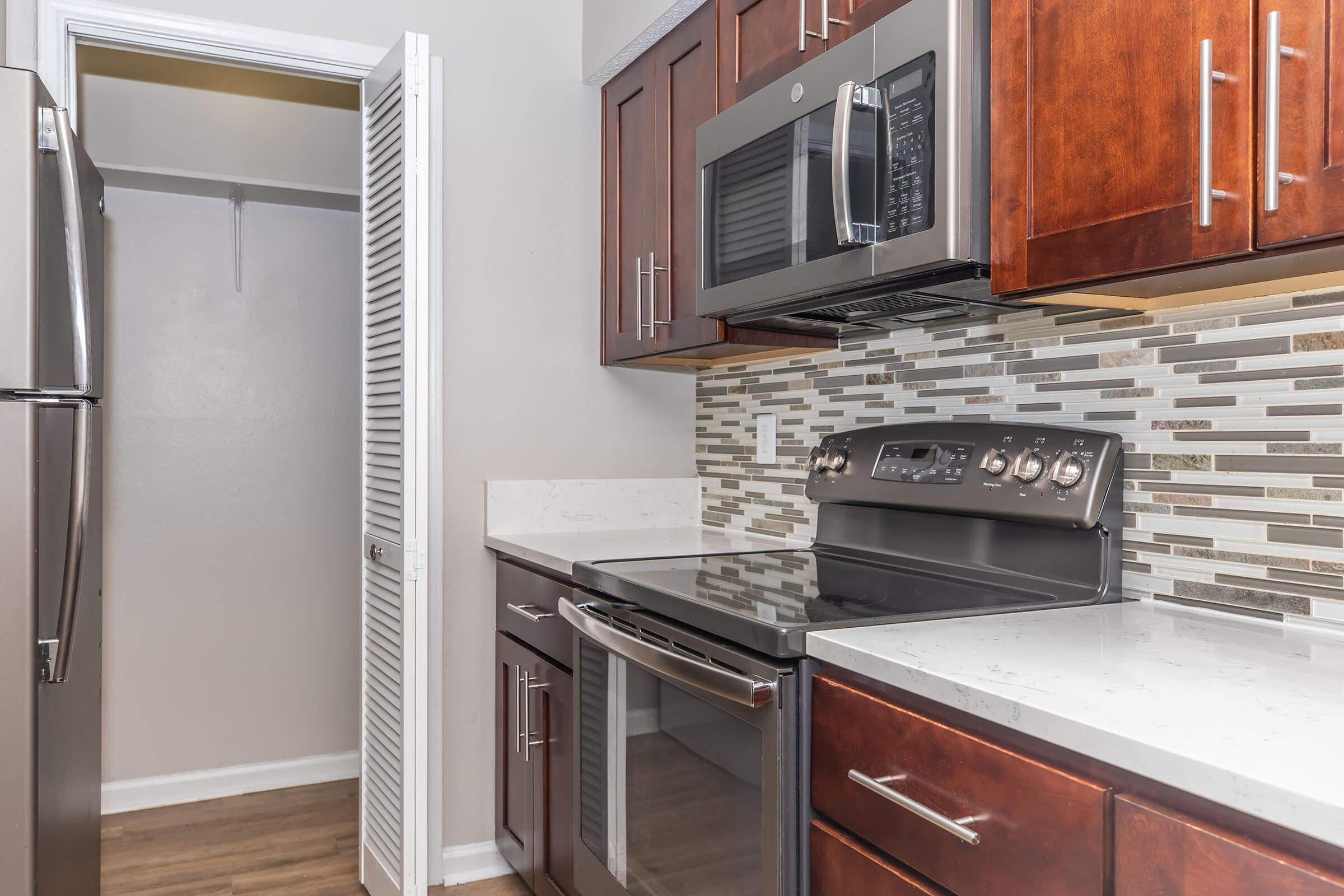 Modern kitchen with dark wooden cabinets, a stainless steel microwave, and a black oven. The countertop is light-colored, contrasting with the cabinetry. A backsplash features a mix of gray and beige tiles. A doorway to a closet or pantry is visible on the left side of the image.