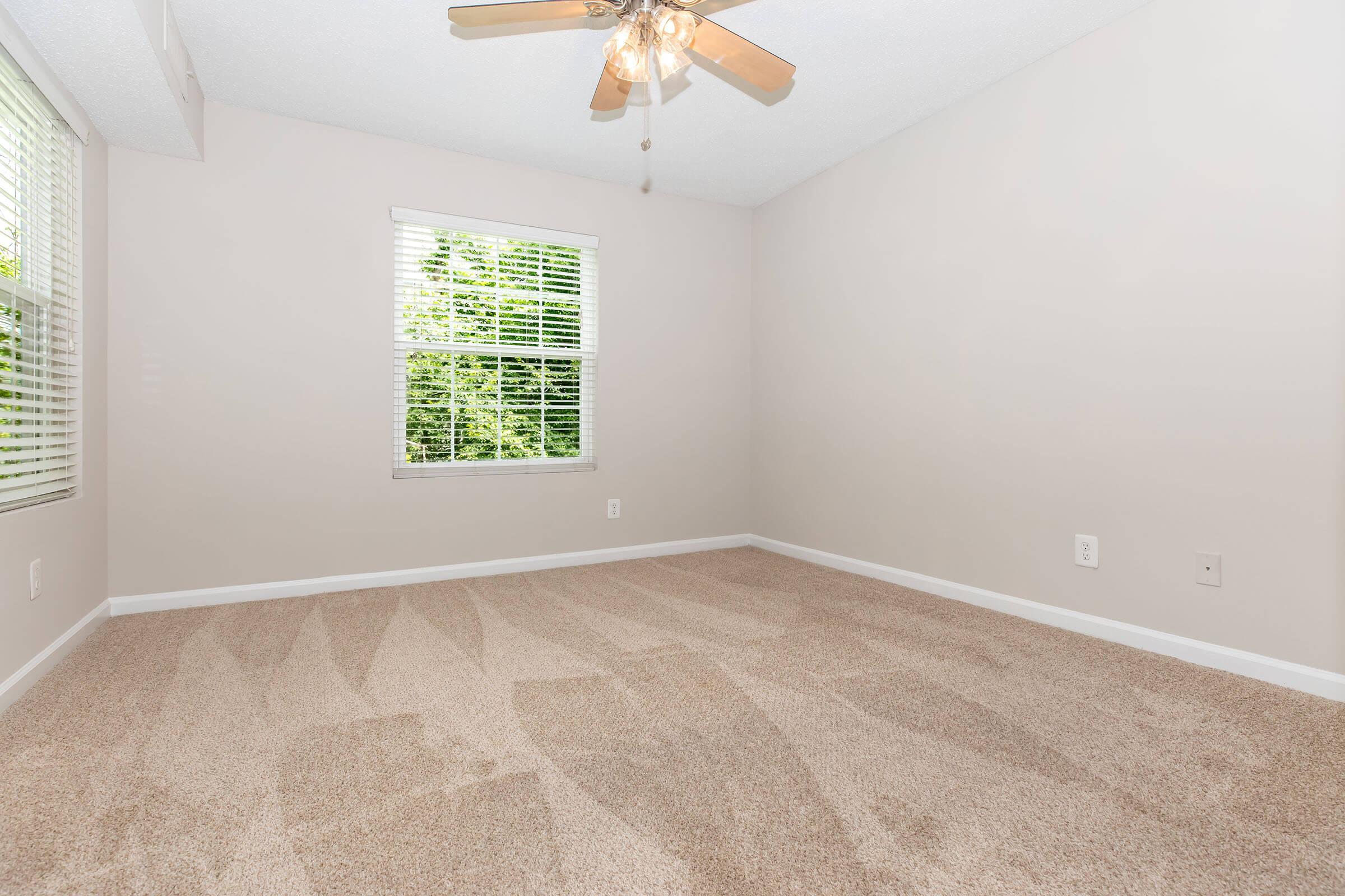 A well-lit, empty room featuring beige carpet and light gray walls. Two windows with white blinds allow natural light to enter. A ceiling fan with wooden blades is installed overhead. The overall atmosphere is spacious and ready for furnishing.