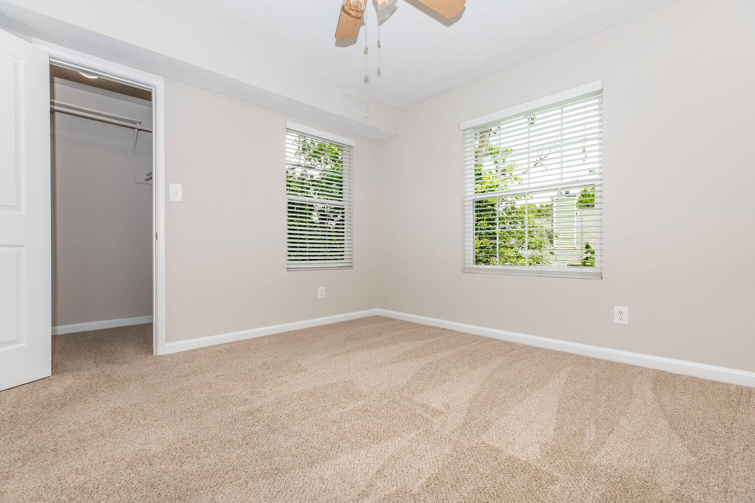 A clean, empty bedroom featuring beige carpet and light gray walls. Two windows with blinds let in natural light, and a ceiling fan is visible. A closet is partially open on the left side. The space appears bright and welcoming, ideal for personal customization.