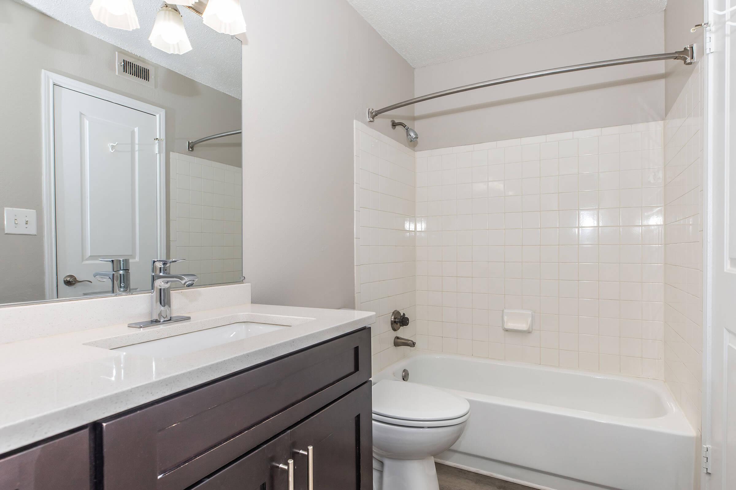A modern bathroom featuring a white bathtub and shower combo, a sleek dark vanity, and a mirror. The walls are painted a light gray, and there is a light fixture with three bulbs above the sink. The floor is tiled in neutral colors, creating a clean and contemporary look.