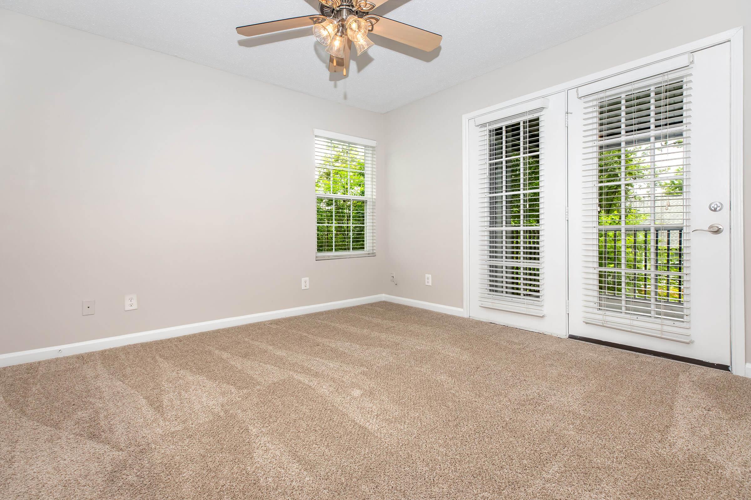 A bright, empty room with light beige carpet and a ceiling fan. Two windows with white blinds let in natural light, and a set of double doors leads to a balcony. The walls are painted a soft gray, creating a calm atmosphere. The space is ideal for various furniture arrangements.