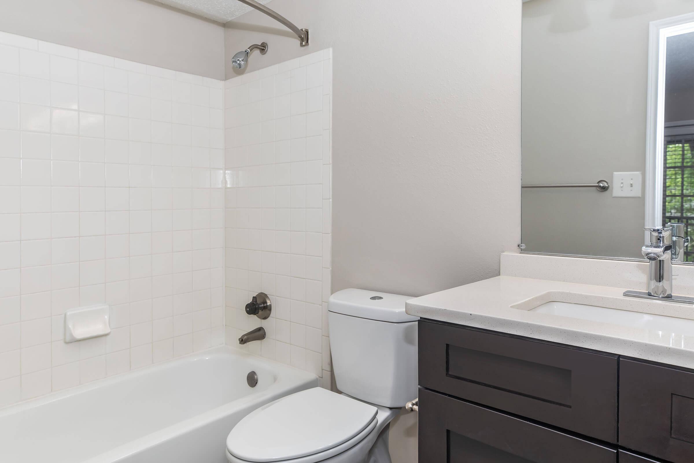 A clean bathroom featuring a white tiled shower and bathtub combination, a modern dark wood vanity with a white countertop, and a toilet. Natural light is coming from a nearby window, creating a bright and inviting space.