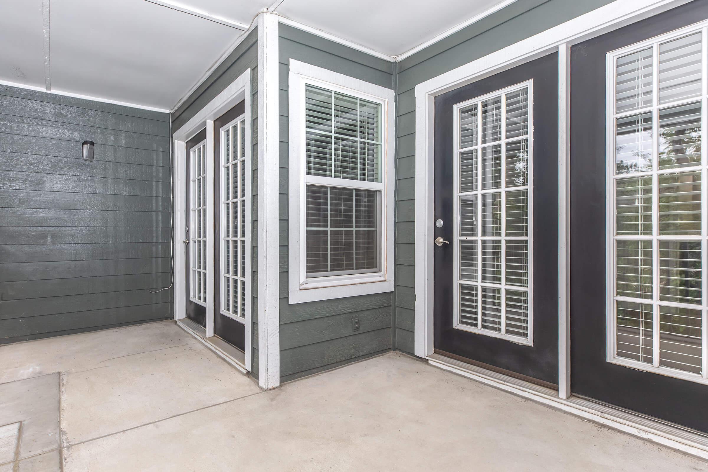 A covered entryway featuring two sets of double doors with glass panels, flanked by a window. The walls are painted a dark green, and the flooring is concrete. The area appears well-lit and enclosed, suggesting a transition from outdoor to indoor space.