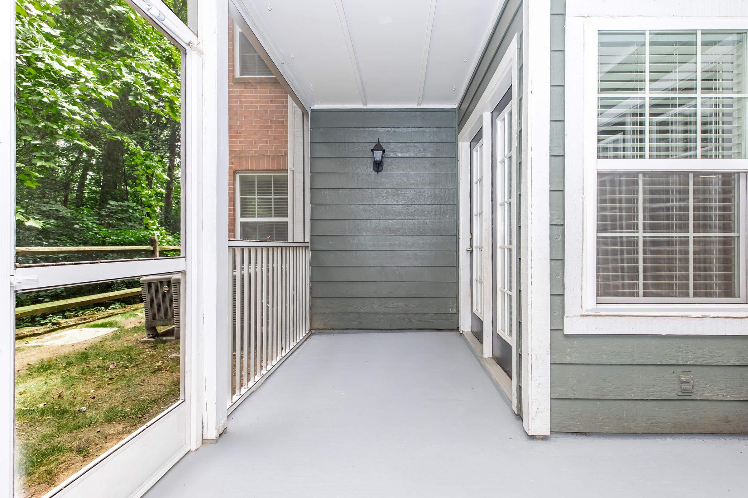 A covered porch area with a light fixture, featuring green and white walls. Large windows provide a view of the outdoors, surrounded by trees. The flooring is light gray, and there is a railing along one side. The space offers a cozy, inviting atmosphere.