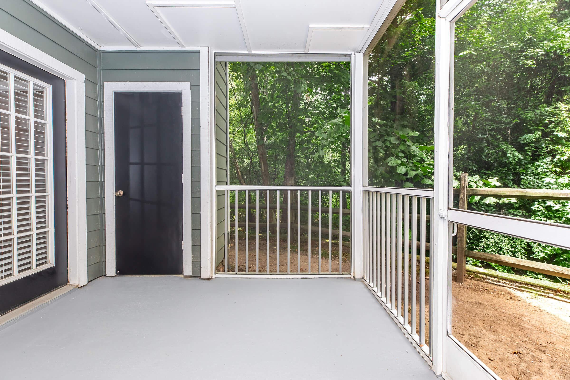A covered porch with a gray floor, white railings, and green surrounding trees visible through screened walls, featuring a black door and a wall with white grid windows. The setting is serene and invites a connection with nature.