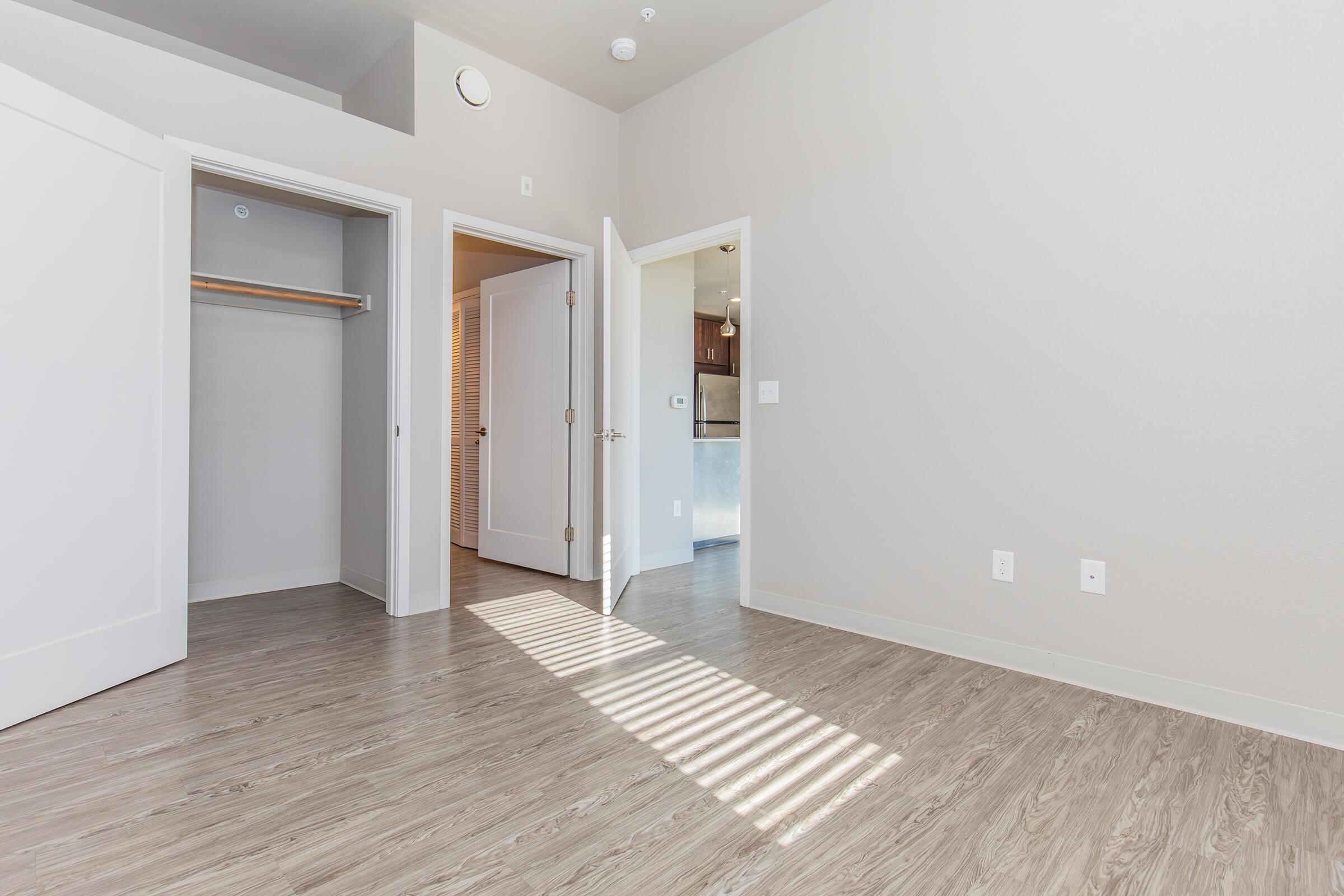 A well-lit interior room featuring light-colored walls and laminate flooring. There are two open doors leading to closets and a bathroom. Natural light filters through the doorways, highlighting the spaciousness of the area.