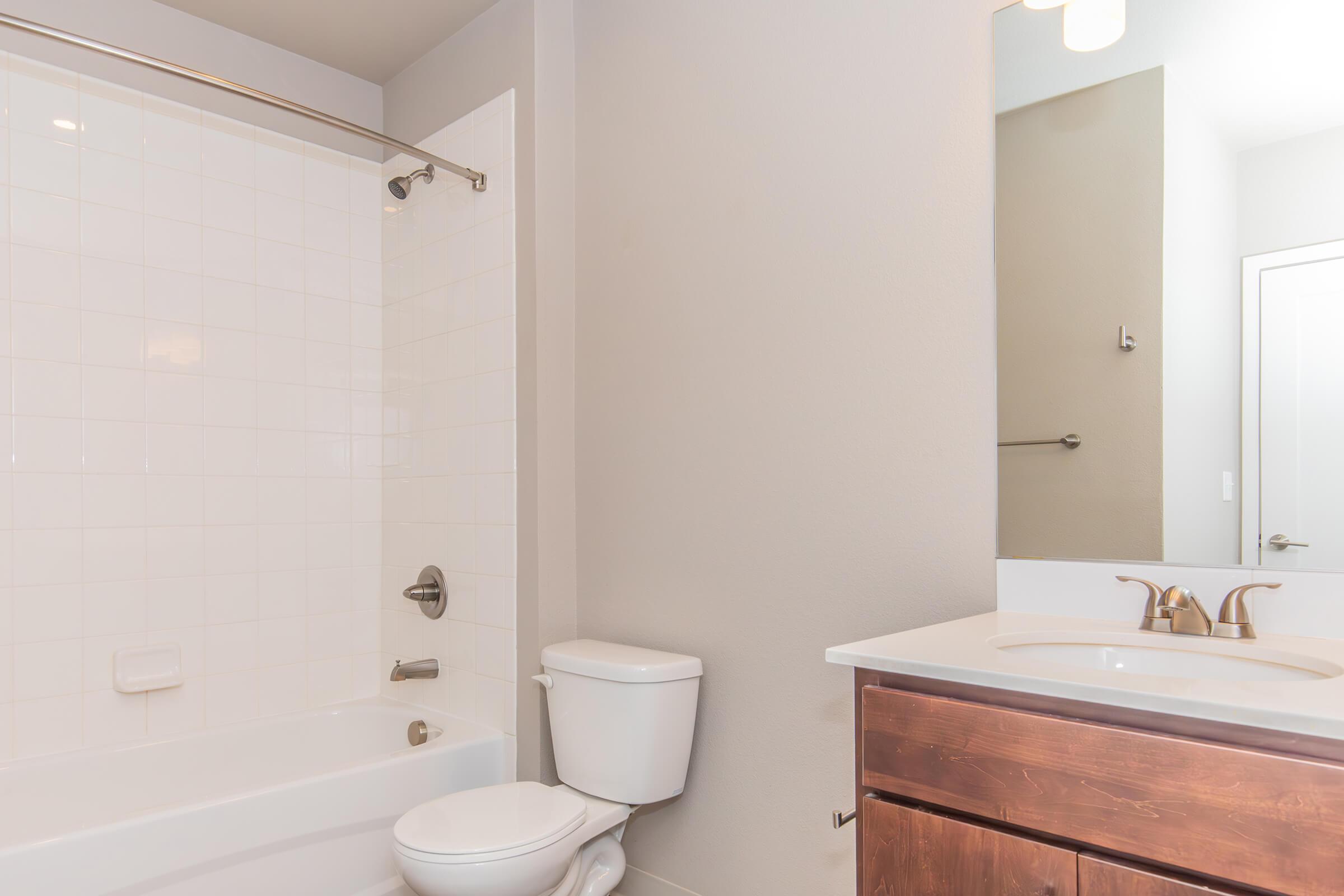 A clean and modern bathroom featuring a white tiled bathtub with a shower, a toilet, and a sleek vanity with a sink and mirror. The walls are painted in a light gray color, and there is a warm light fixture above the mirror. The overall design is simple and contemporary.