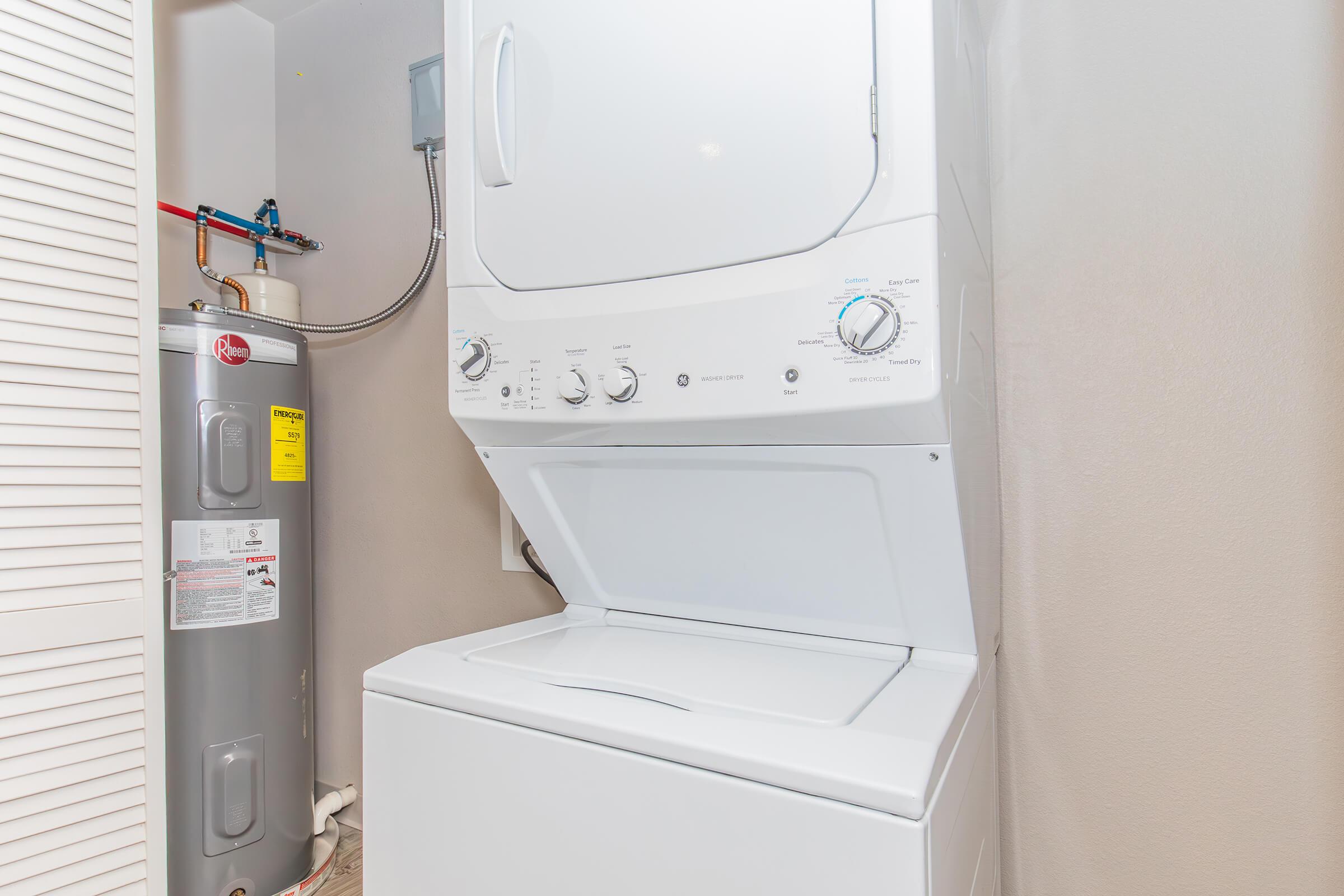 A stackable washer and dryer set in a compact laundry space, next to a water heater. The appliances are white, and the space features light-colored walls and flooring.