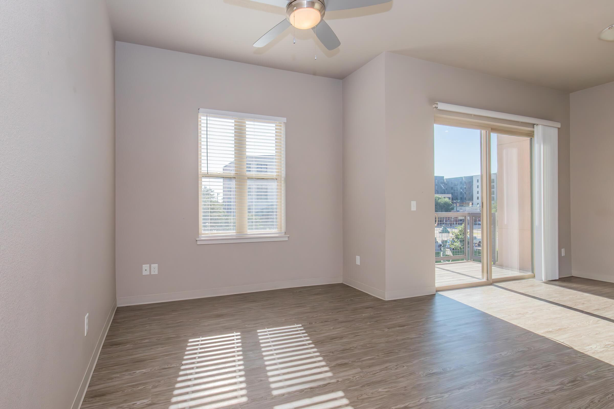 Bright, empty room with light-colored walls and wooden flooring. A large window provides natural light, and there's a door leading to a balcony. A ceiling fan is visible above. The space has a clean and modern feel, ideal for decoration or furnishing. No furniture is present.