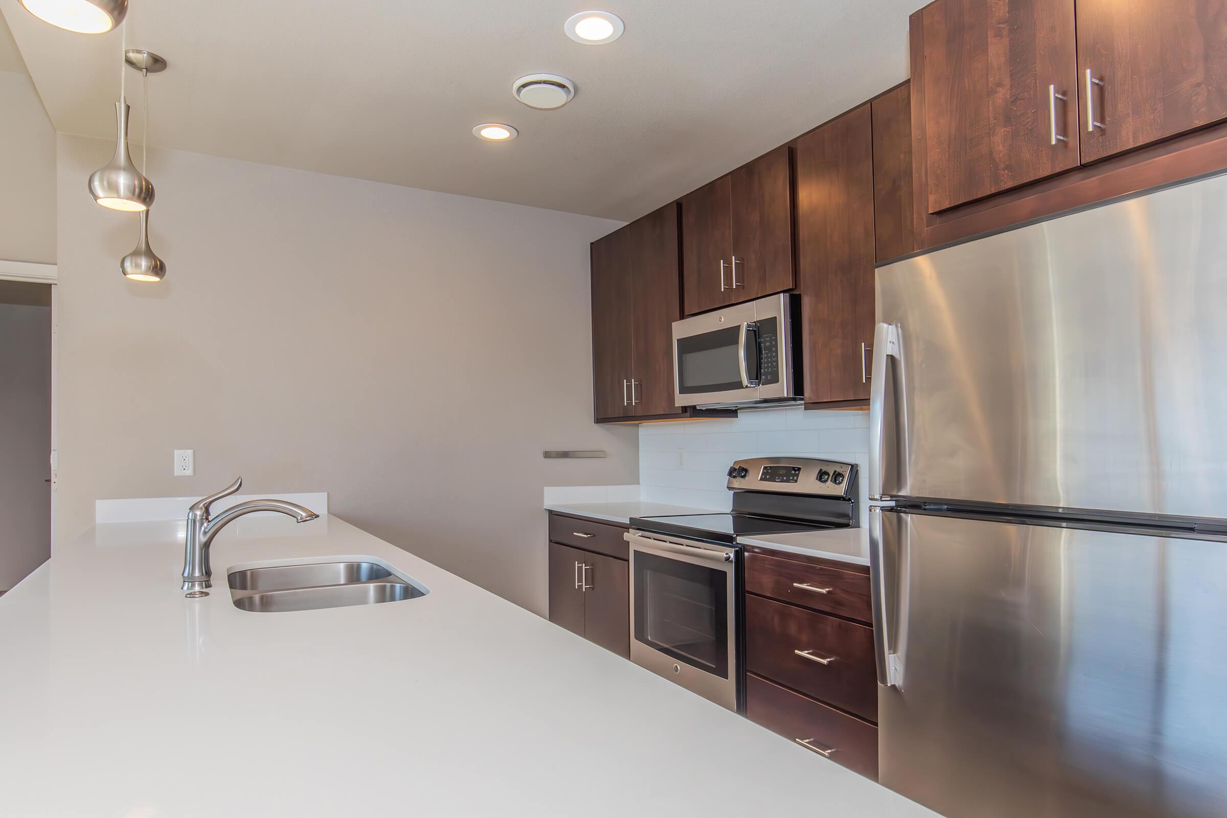Modern kitchen featuring sleek dark wood cabinets, stainless steel appliances including a microwave and refrigerator, and a contemporary countertop with a built-in sink. Soft lighting from pendant fixtures illuminates the space, creating a clean and inviting atmosphere.