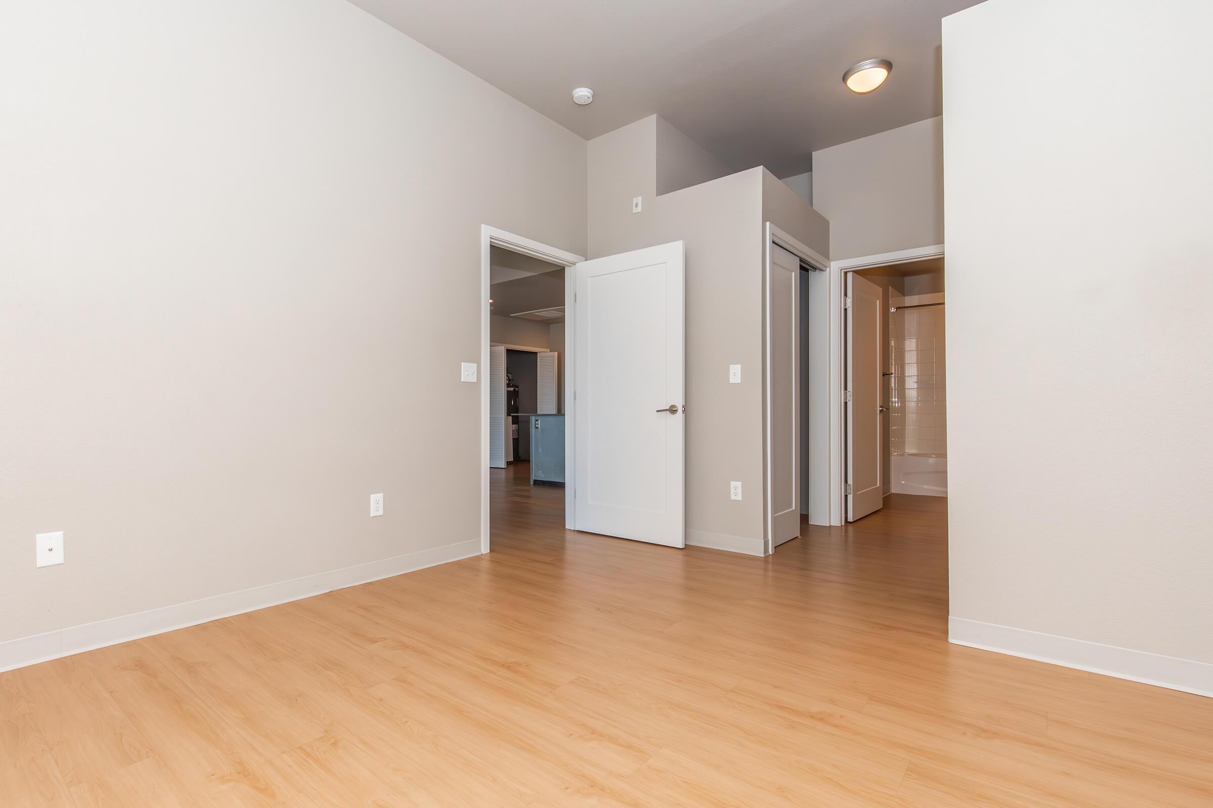 A spacious, well-lit room featuring light wood flooring, beige walls, and three doorways leading to other areas. Natural light enters from an unseen source, enhancing the open feel of the space. There is a simple ceiling light fixture visible in the upper left corner.