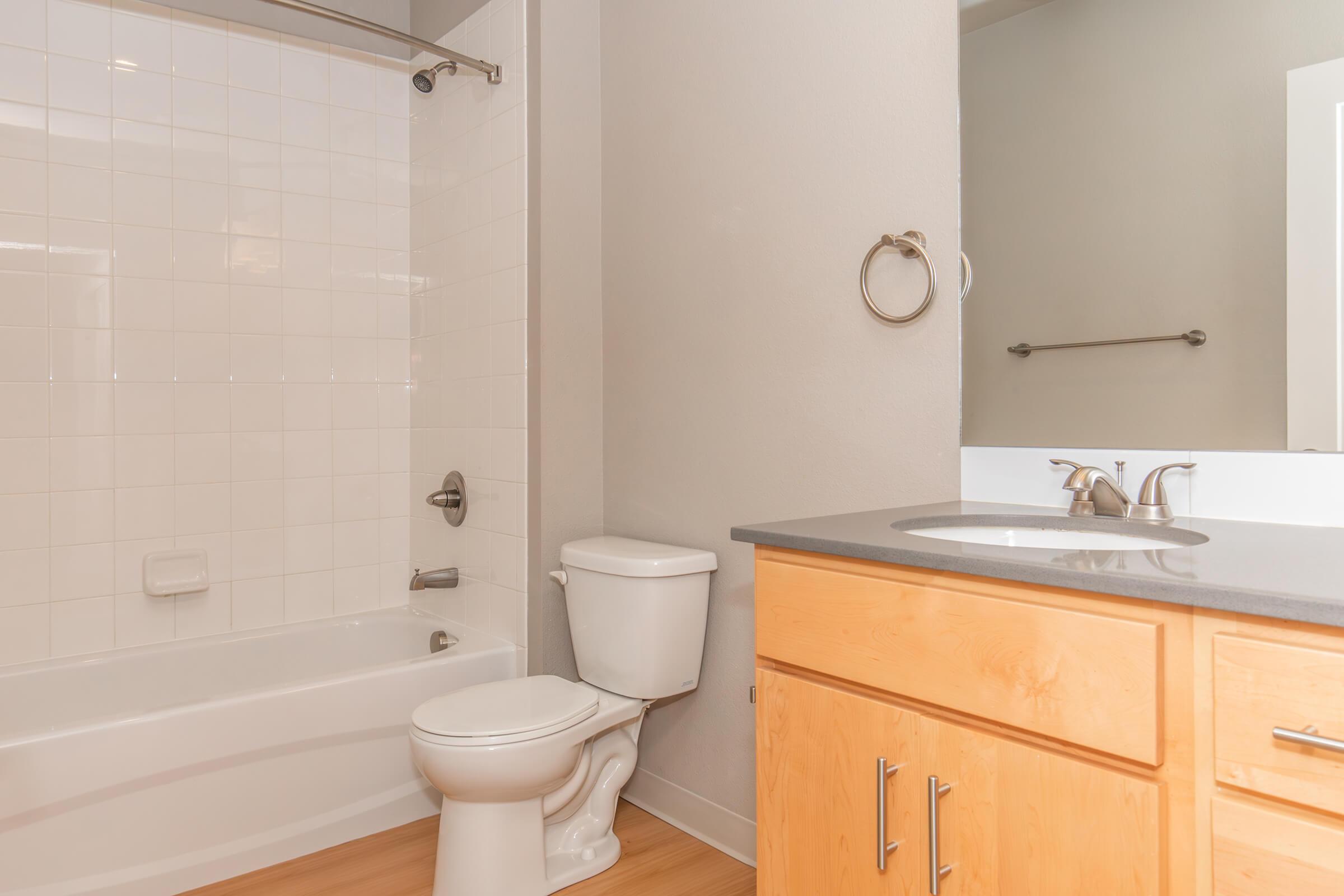 A modern bathroom featuring a white tiled bathtub with shower, a toilet, and a wooden vanity with a sink. The walls are painted in neutral colors, and there is a mirror above the vanity. Light wood flooring complements the clean and simple design.