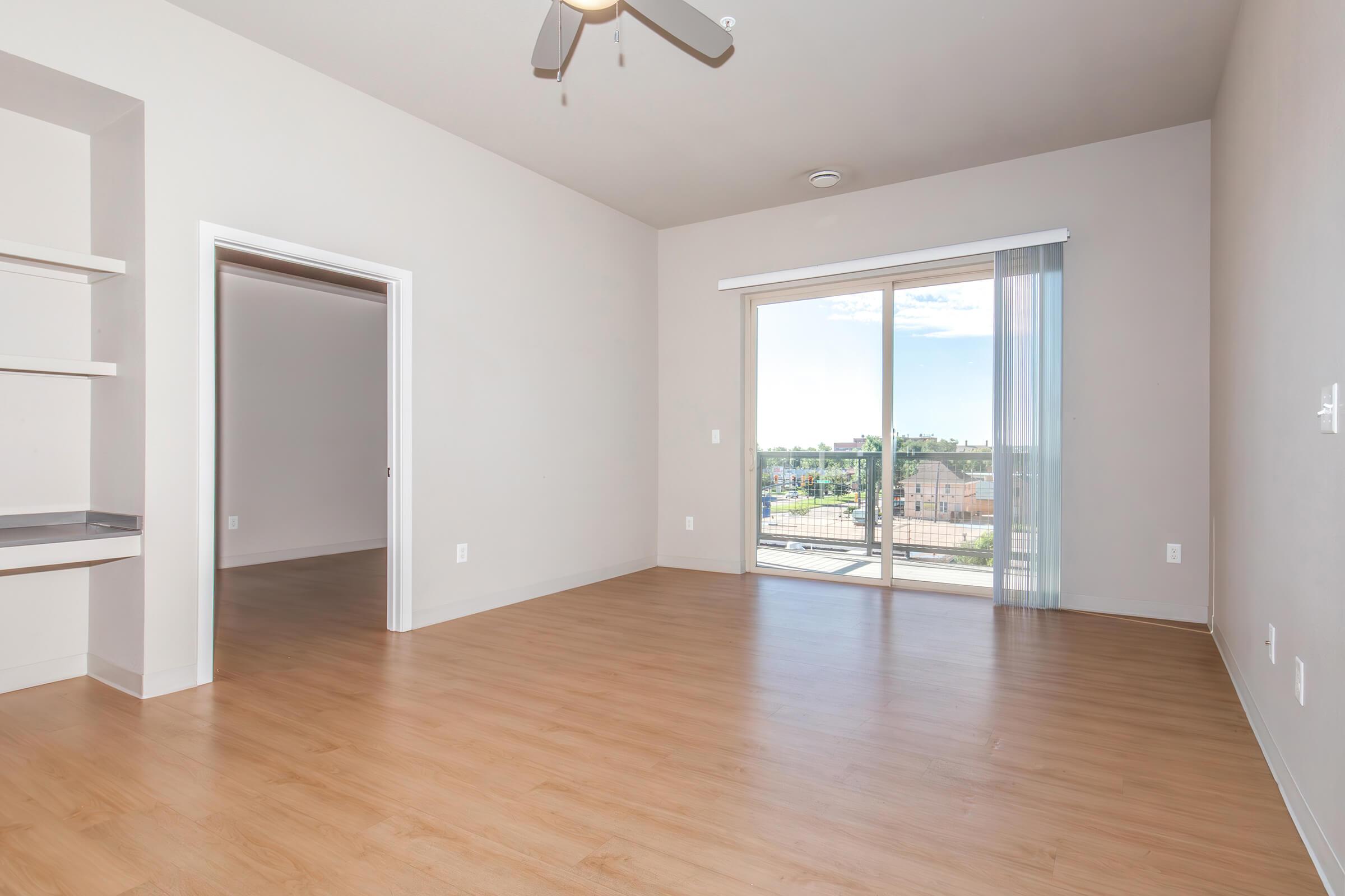 A bright, spacious living room with light-colored walls and wood-like flooring. There is a ceiling fan, a sliding glass door leading to a balcony, and a doorway to another room. Large windows provide ample natural light. The space is unfurnished, creating an open and airy atmosphere.
