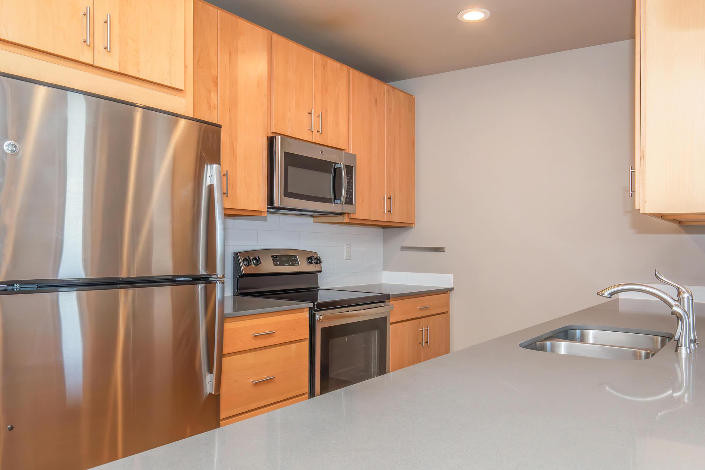 Modern kitchen featuring stainless steel appliances including a refrigerator, microwave, and oven, alongside wooden cabinets and a sleek countertop. A double sink is visible, enhancing the contemporary design of the space. Natural light illuminates the area, creating an inviting atmosphere.