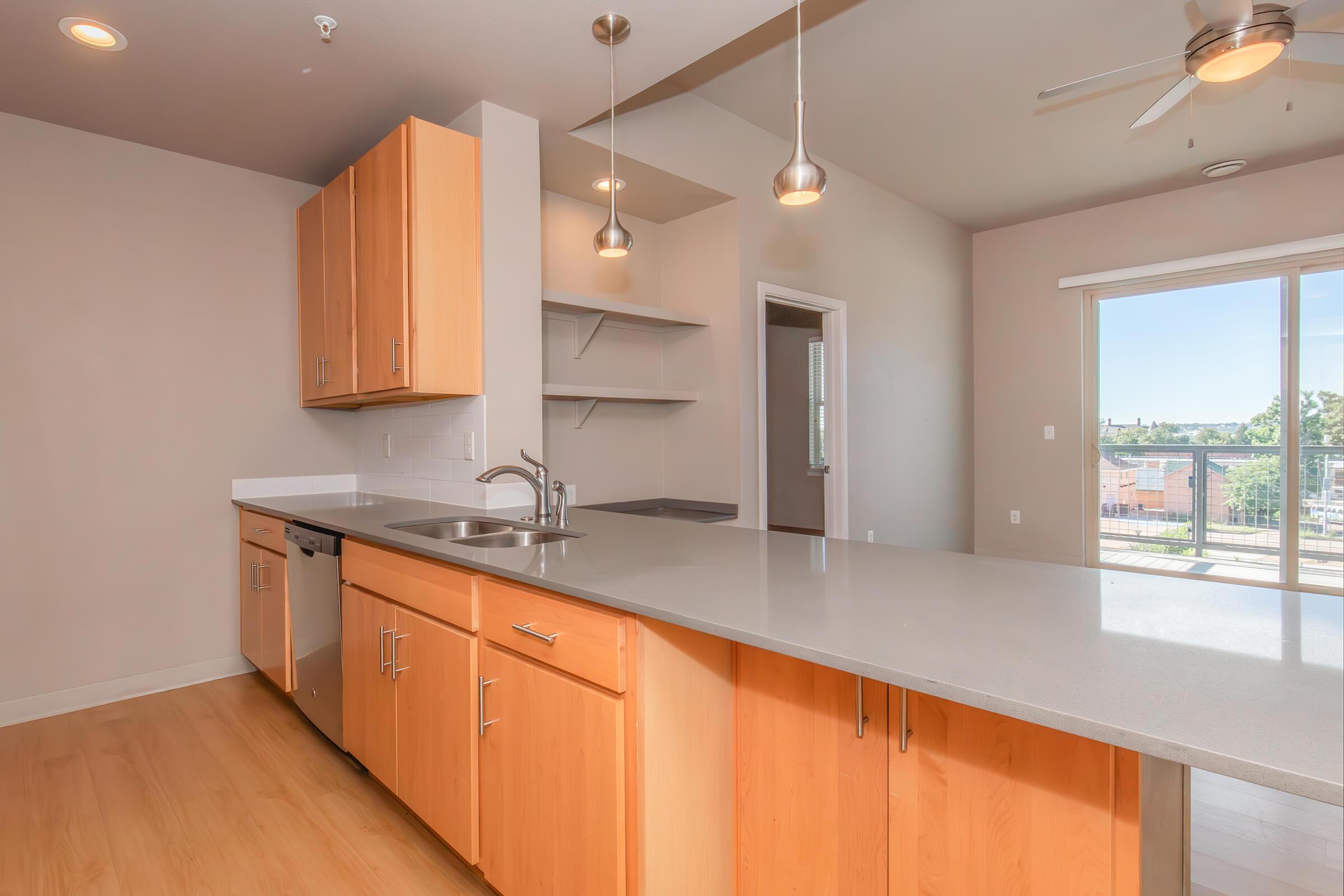 Modern kitchen featuring light wood cabinetry, stainless steel appliances, and a large countertop island. The room has a neutral wall color and ample natural light from a large sliding glass door leading to a balcony. Shelving units are visible in the background.