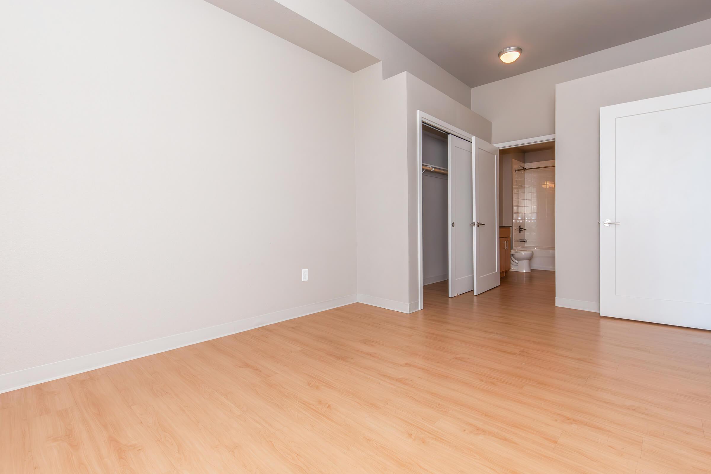 A spacious, empty room with light-colored walls and laminate flooring. The room features a closet on the left and an open door leading to a bathroom beyond. Natural light brightens the space, highlighting the clean, minimalist design.