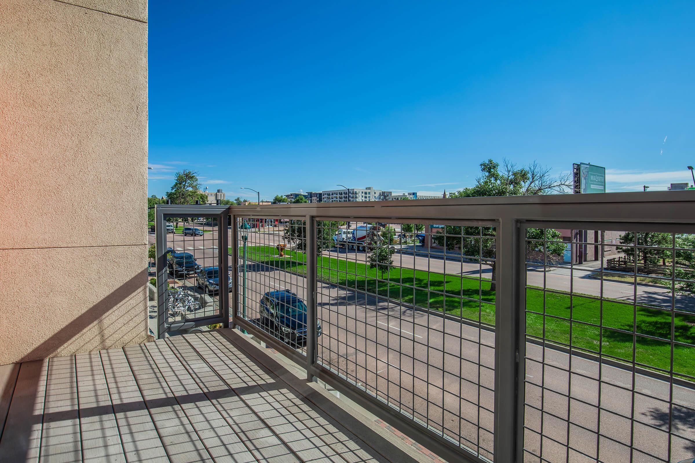 A balcony view overlooking a street with parked cars and grassy areas. The sky is clear blue, and the area is well-lit by sunlight, highlighting the railing's design and the surrounding greenery.