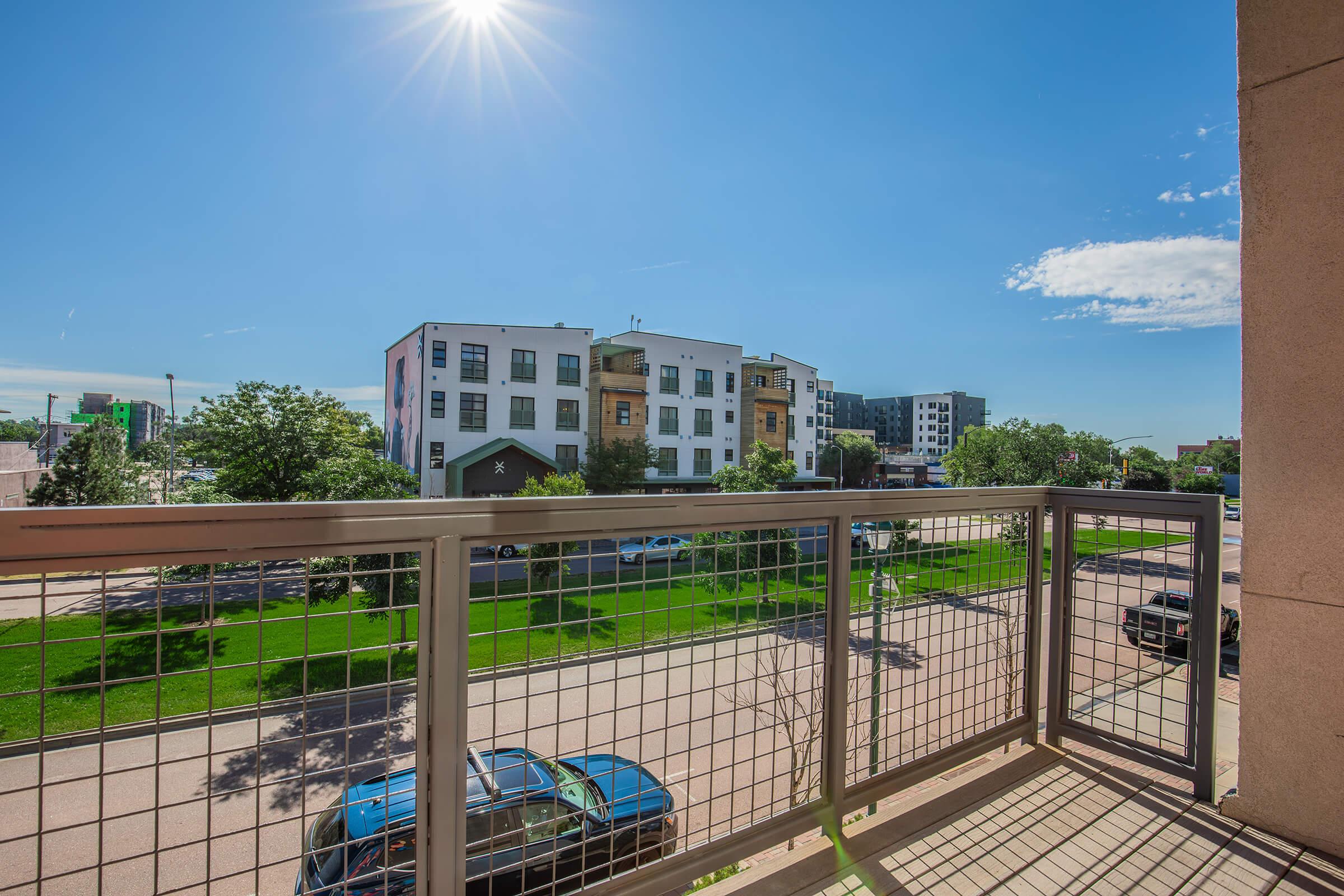View from a balcony showcasing a clear blue sky and bright sun. In the foreground, there is a modern building with several apartments, surrounded by greenery and trees. Below, a street with parked cars and pedestrian pathways can be seen, contributing to a lively urban atmosphere.