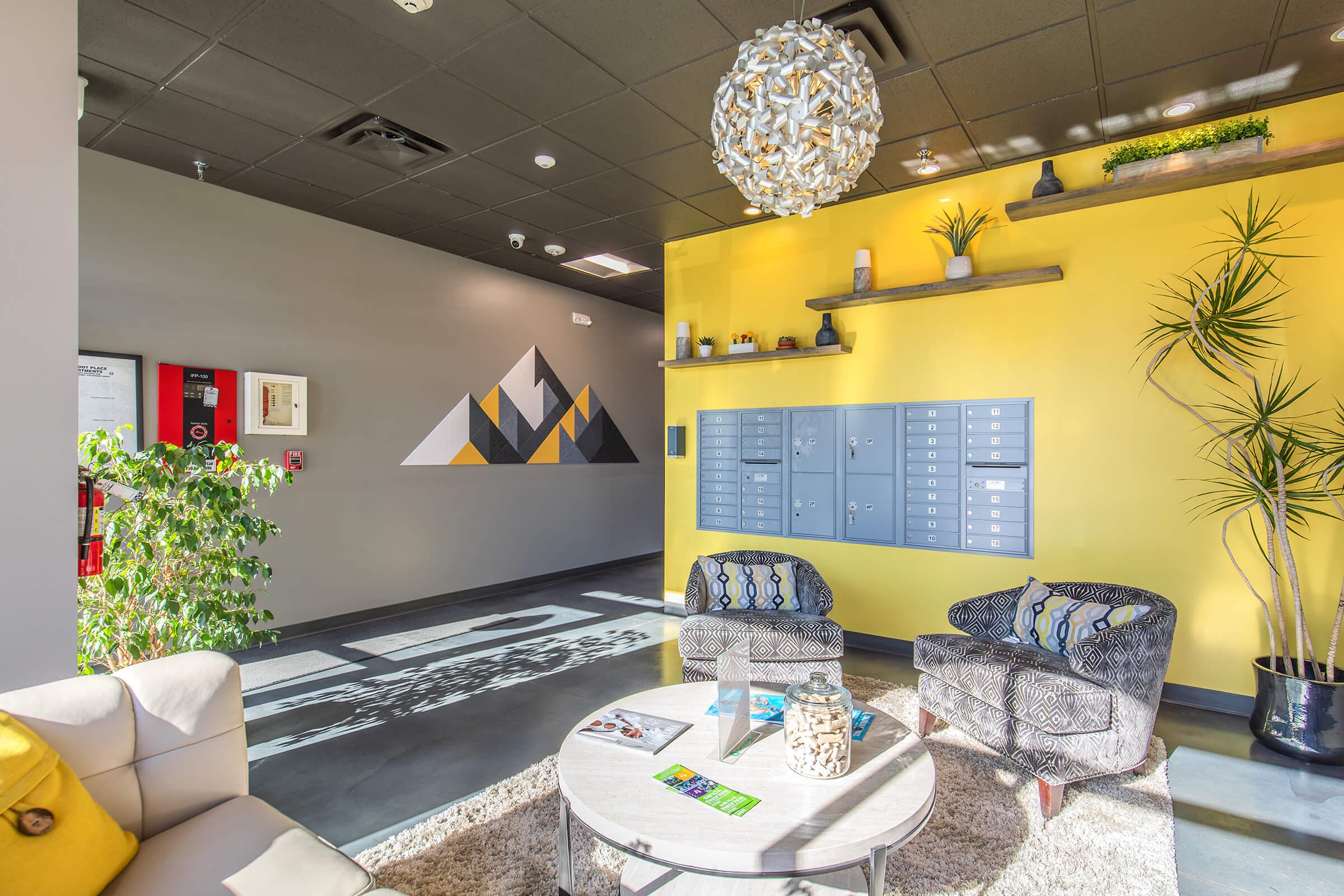Modern interior of a lobby featuring a yellow accent wall with a geometric mural, gray wall with mailboxes, two stylish chairs with patterned cushions, a round coffee table with decorative items, and natural plants. Bright lighting and sleek design elements create a welcoming atmosphere.