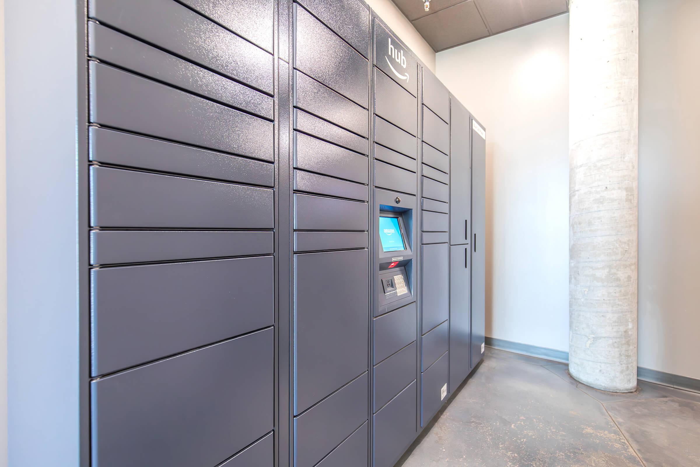 A series of gray delivery lockers with a digital control panel in front, set in a modern indoor space with concrete flooring and walls. The lockers are organized in a row against a wall, offering a convenient solution for package pickup and storage.