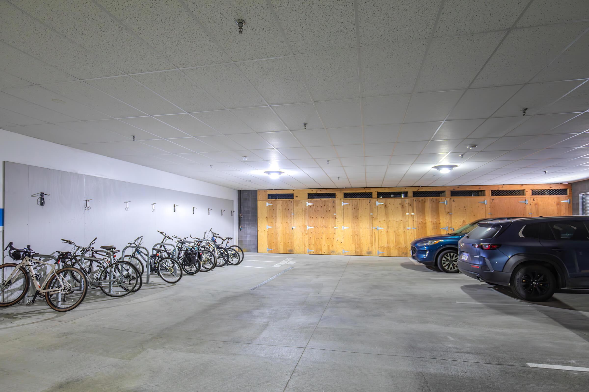 A spacious indoor parking area featuring several bicycles lined up against a wall, and two parked vehicles—one blue SUV and another with a darker tone. The walls are painted light gray, and wooden panels cover a section of the back wall, providing a neat appearance to the space. Bright ceiling lights illuminate the area.