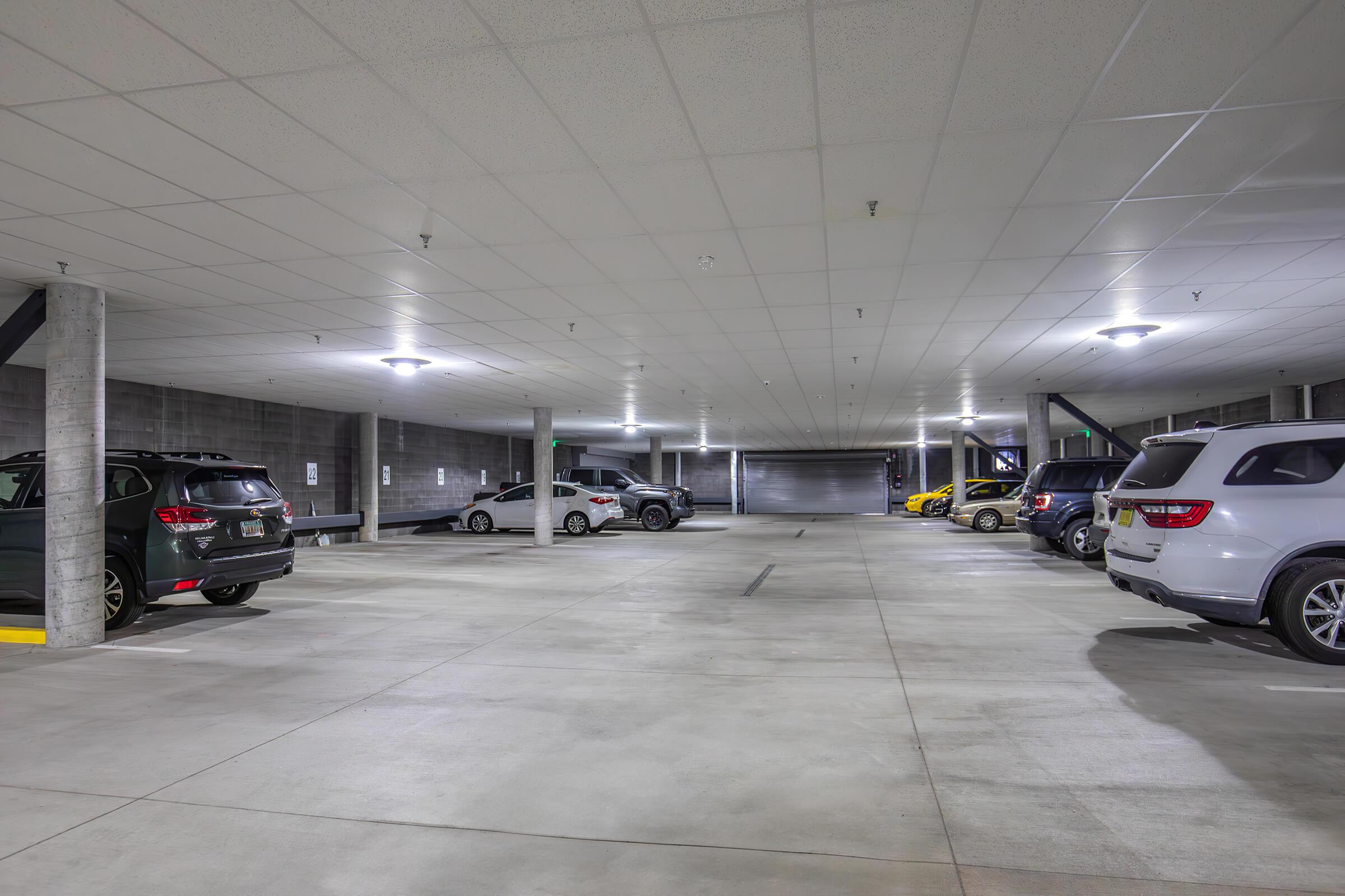 A spacious, well-lit underground parking garage featuring parked cars in various colors. The ceiling is white with overhead lights, and there are concrete pillars evenly spaced throughout the area. The ground is smooth with marked parking spaces.