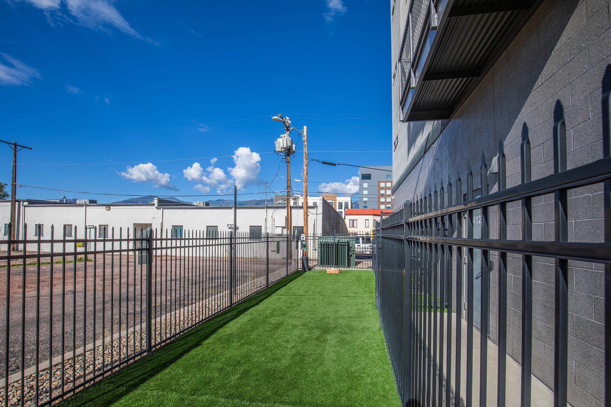 A view of a secluded outdoor area with artificial grass, bordered by a black metal fence. In the background, there are utility poles and buildings under a bright blue sky with scattered clouds. The setting appears clean and well-maintained, offering a quiet space.