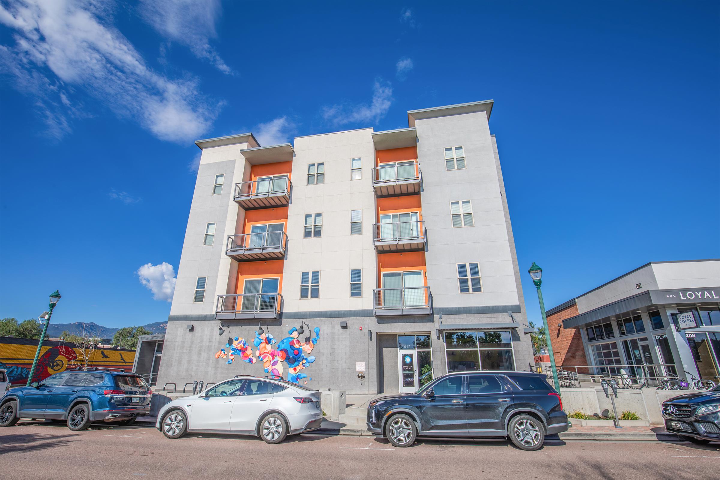 Modern multi-story building with a colorful mural on the ground floor. The structure features multiple balconies and large windows, situated on a lively street with parked cars. Bright blue sky and a few clouds enhance the urban scene. Nearby buildings can be seen in the background.