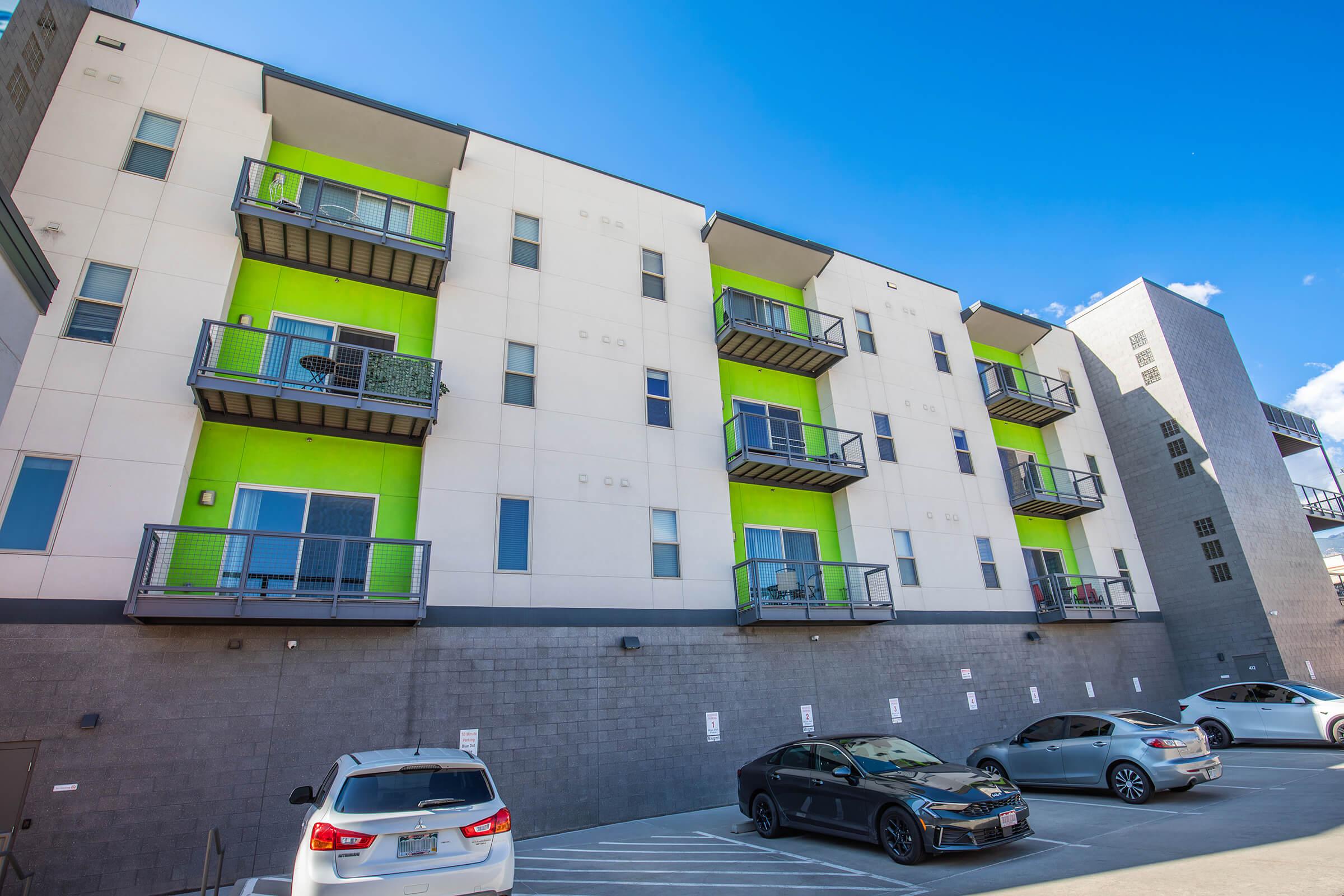 A modern multi-story apartment building featuring a white and green exterior with multiple balconies. Several cars are parked in front of the building, and the sky is clear and blue.