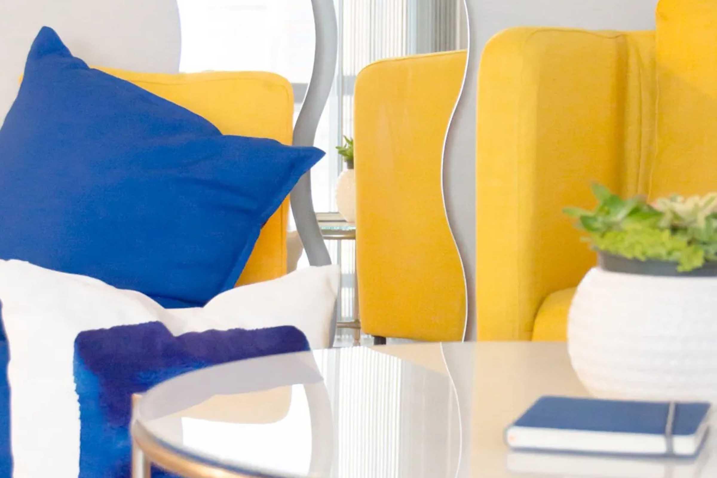 A bright living space featuring a yellow armchair and a blue cushion on a white sofa. A round glass coffee table reflects the light, with a white pot containing a small plant. A blue notebook rests on the table, adding to the contemporary decor. The overall atmosphere is cheerful and inviting.