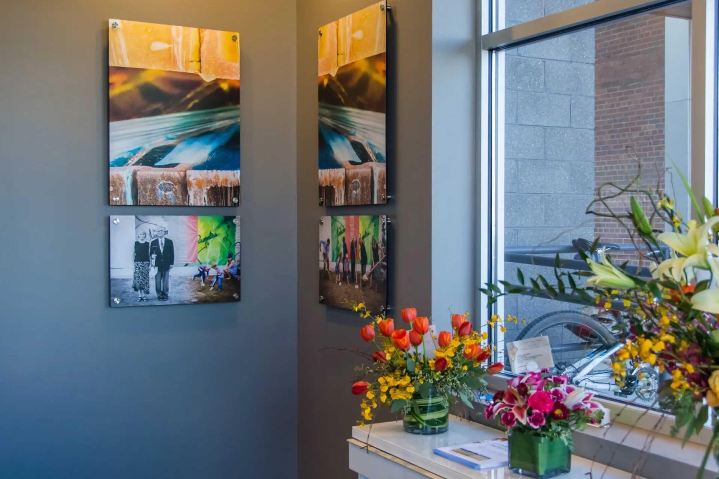 A corner of a gallery or waiting area featuring colorful artwork displayed on gray walls. The images include abstract designs and black-and-white photographs of people. A table adorned with vibrant flower arrangements and a few cards is in the foreground, adding a touch of nature and color to the contemporary space.