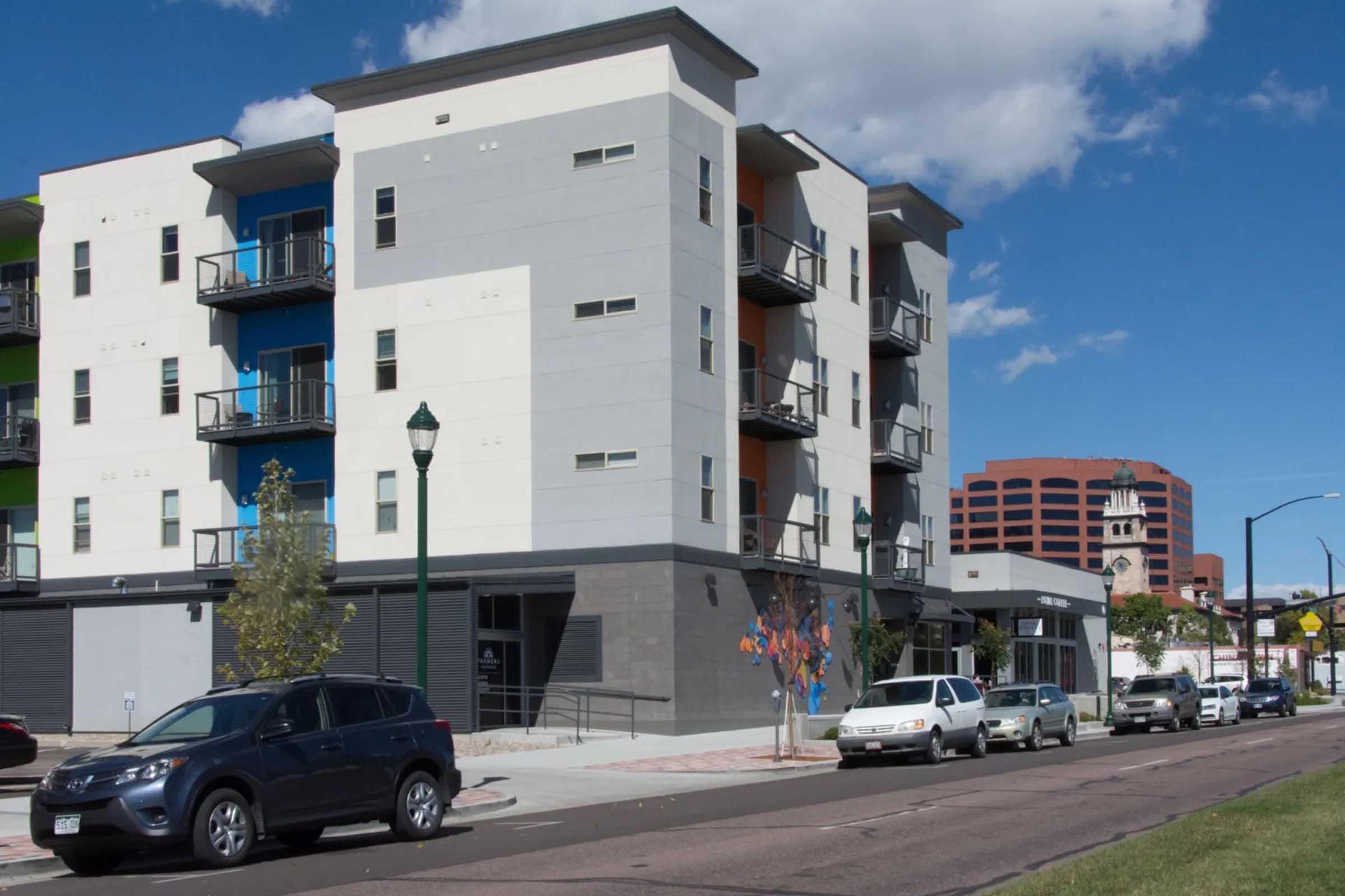 Modern multi-story residential buildings with colorful facades. The structure features balconies and large windows. In the foreground, there are parked cars along a sidewalk, with trees and streetlights visible. Clear blue sky with some clouds adds to the urban setting.