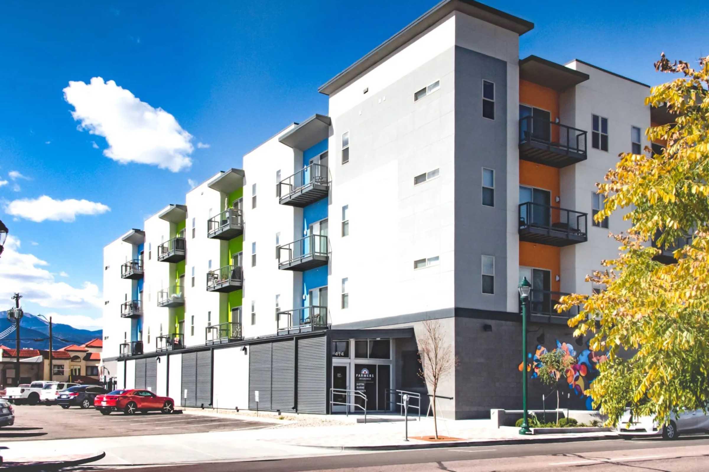 Modern multi-story apartment building featuring a mix of light and dark exterior colors, with balconies on each floor. The ground level contains storefronts, and there are parked cars in the foreground. Bright blue sky with a few clouds is visible in the background.