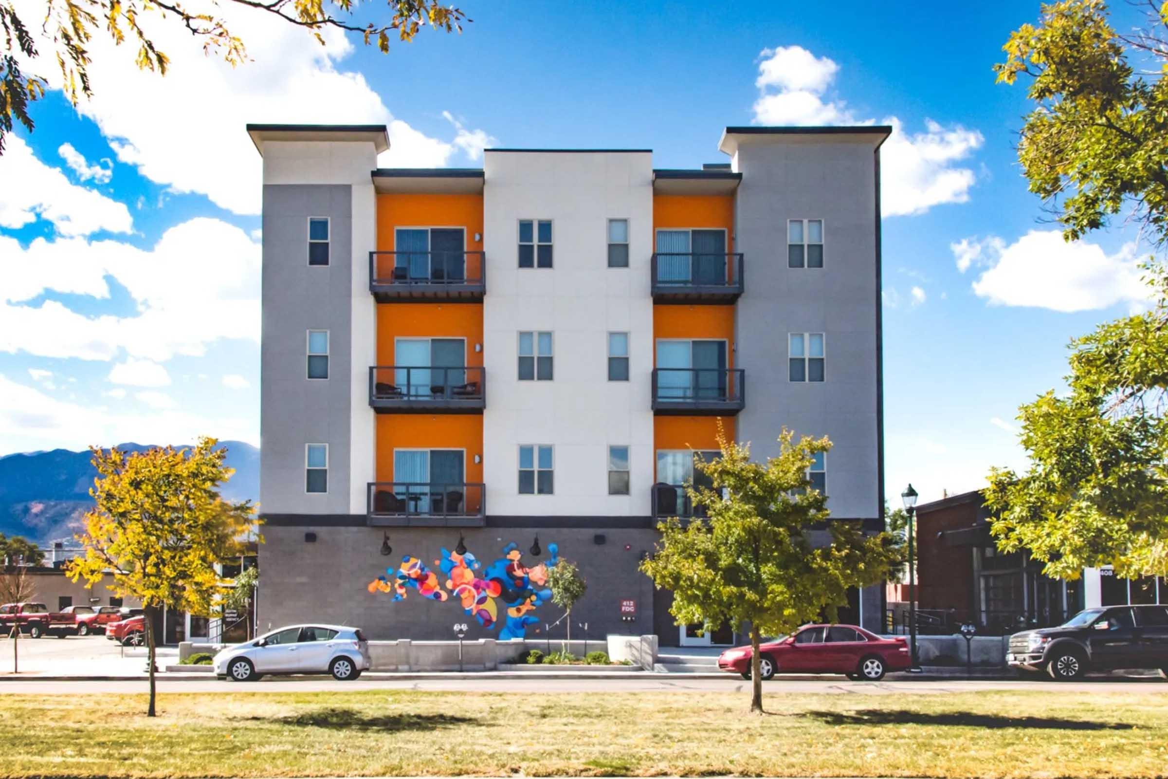A modern four-story apartment building with orange and gray exterior, featuring balconies on each floor. In front, there is a colorful mural on the wall and parked cars along the street. Surrounding trees and a grassy area add to the vibrant scene under a blue sky with scattered clouds.